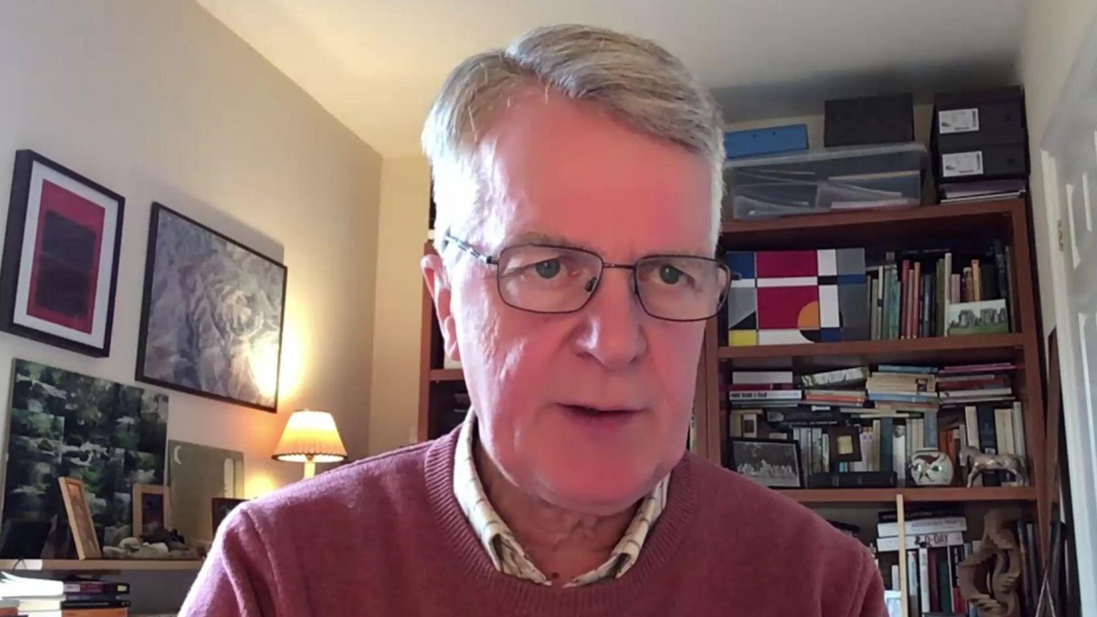 John Adams wears glasses on an online video call, with bookshelves in the background and wearing a red jumper