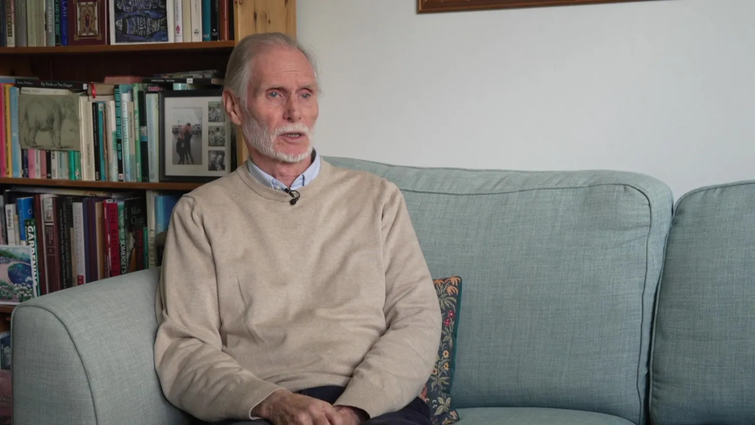 Jim with white hair and a white beard, wearing a beige jumper over a blue collared shirt, sits on a pale green sofa in front of a full bookcase as he speaks during an interview