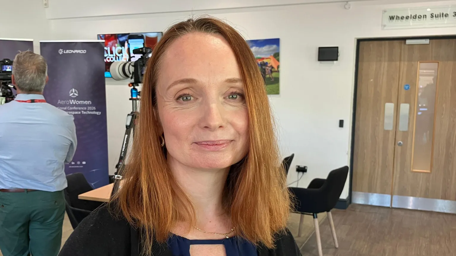 A woman is stood in the middle of the frame, smiling at the camera. She has shoulder length red hair. She is wearing a black cardigan and blue shirt. Behind her is a man filming someone on a camera.