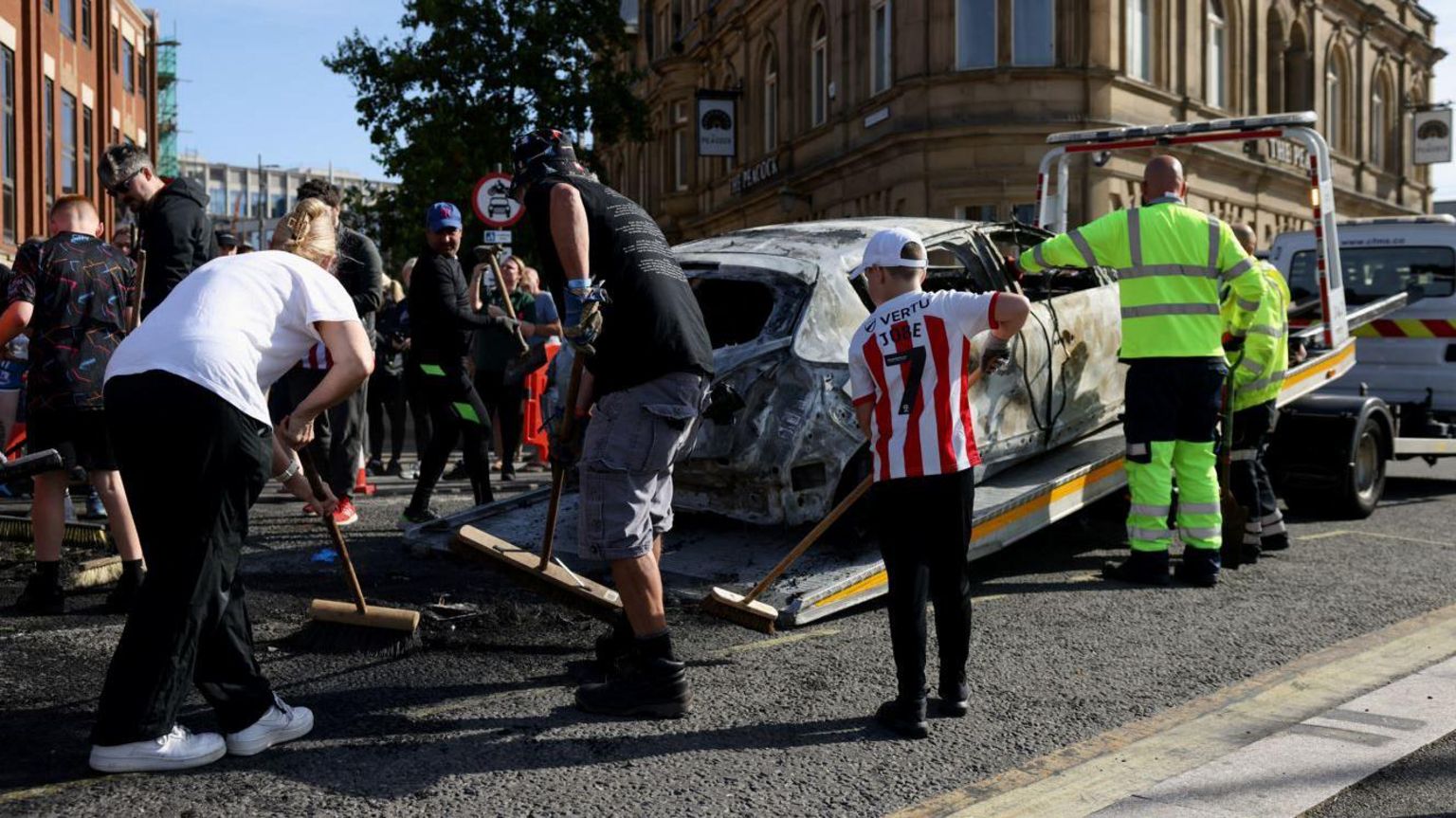 Sunderland residents using brooms to clean the city after the riot. A boy wearing a Sunderland football shirt is among those sweeping. A burnt-out car is being loaded onto the back of a van to be removed.