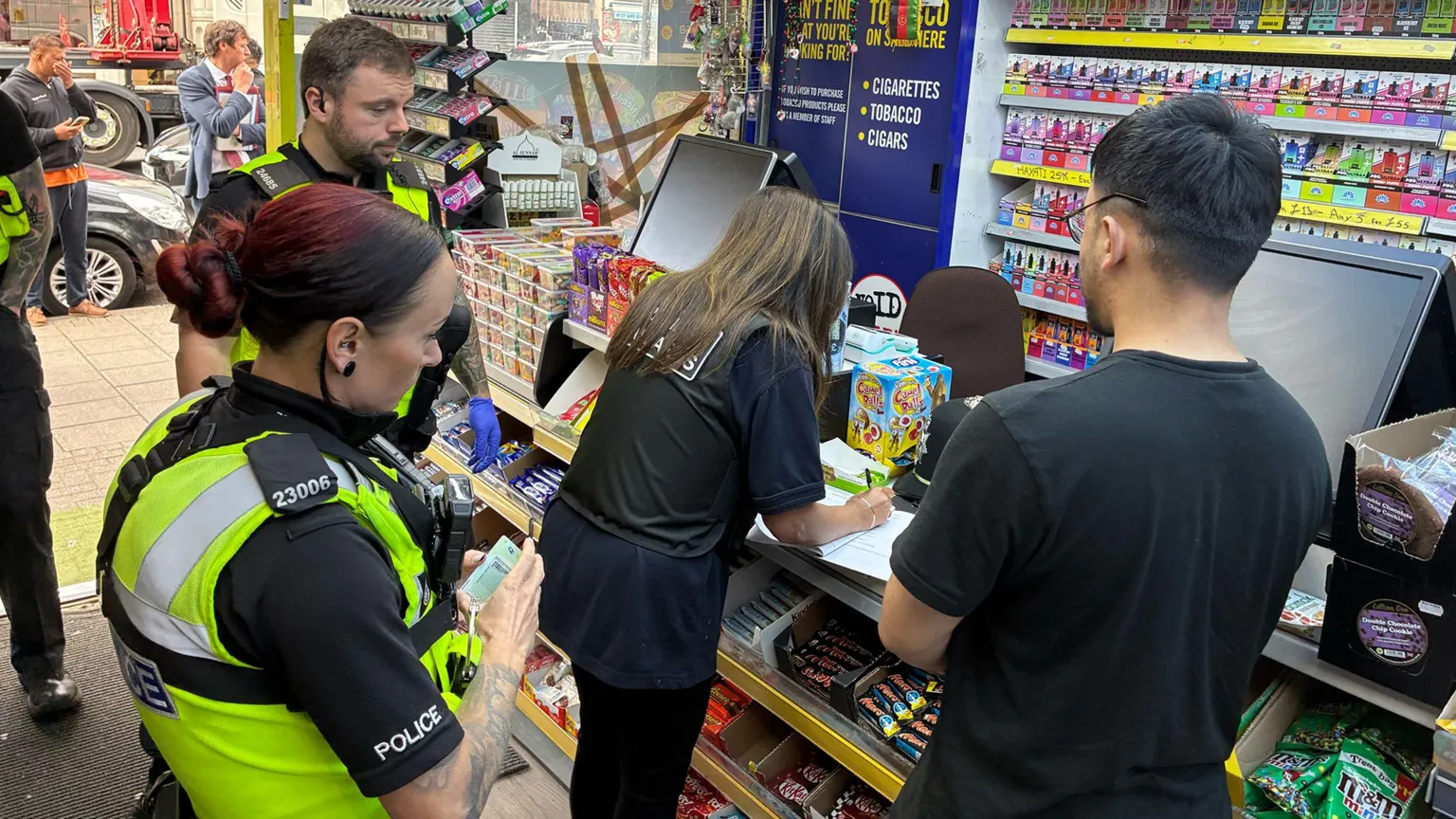 Police officers stand inside a convenience store as a shop worker and Trading standards official fill out paperwork at the counter, with shelves of sweets, cigarettes and colourful vaping products behind them.