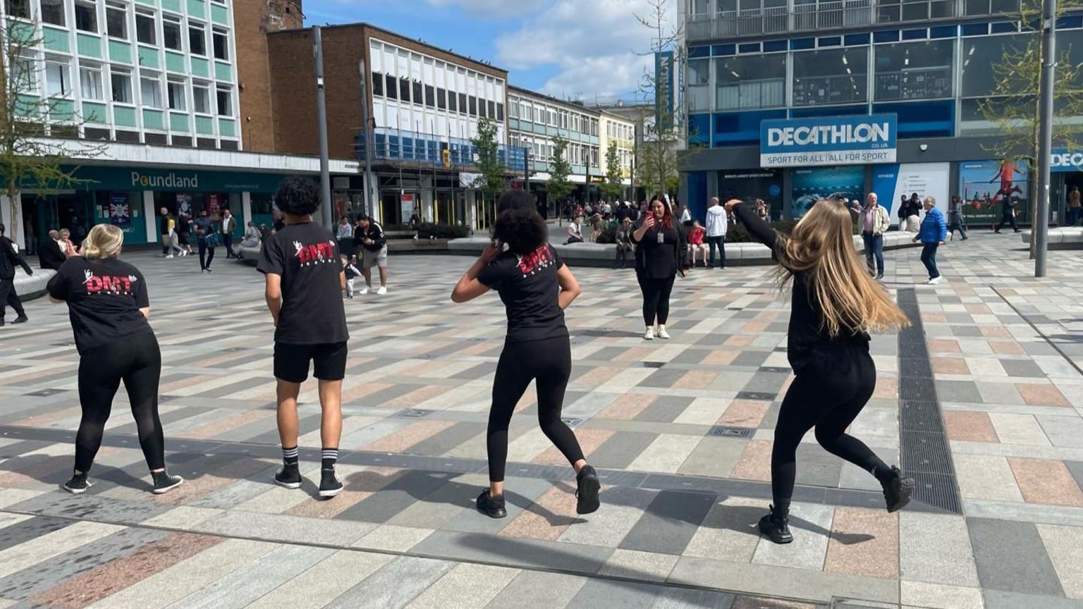 Four young dancers facing away from camera perform dressed all in black in the centre of Crawley town, with shops an spectators surrounding them