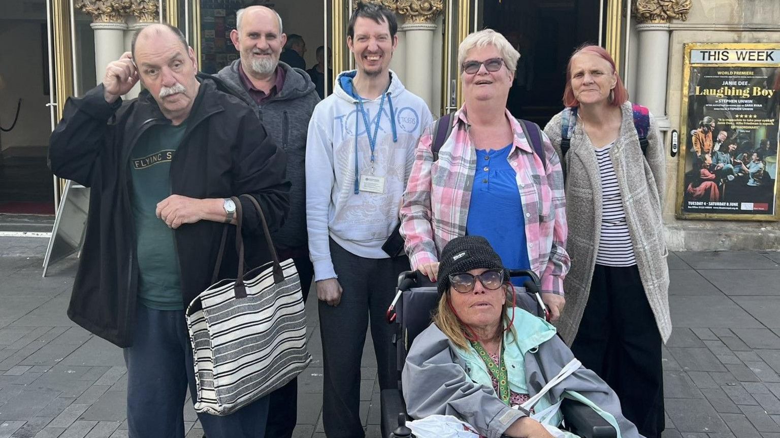 Michael Edwards in front of theatre for the Laughing Boy play. He is the first of six people from left to right. The five other people are beside him, one is a wheelchair user. They are all facing the camera. In the background, there is a poster for Laughing Boy that says the play is on this week.