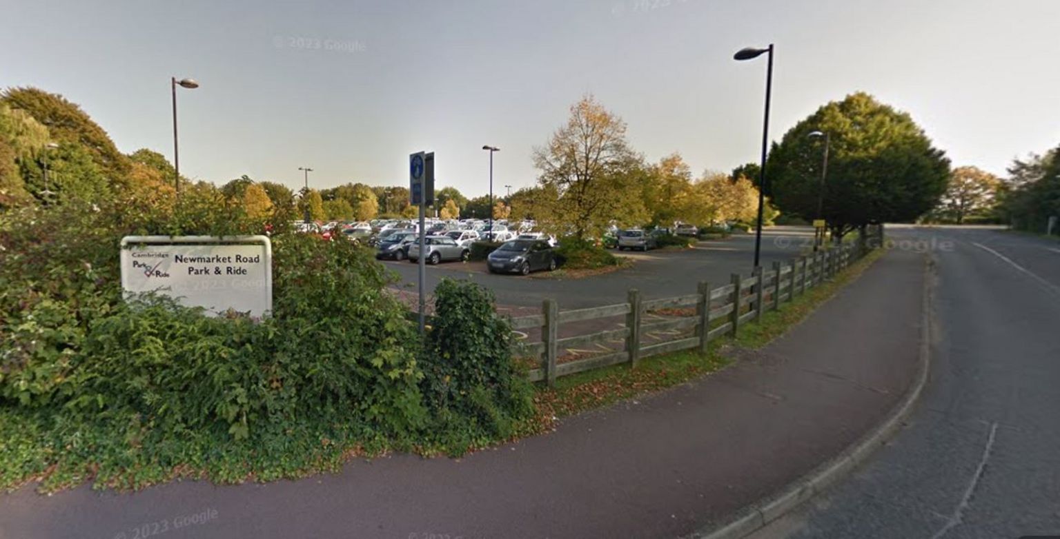 Sign which says Newmarket Road Park and Ride surrounded by bushes. A wooden fence is to the right and cars are parked behind it. 