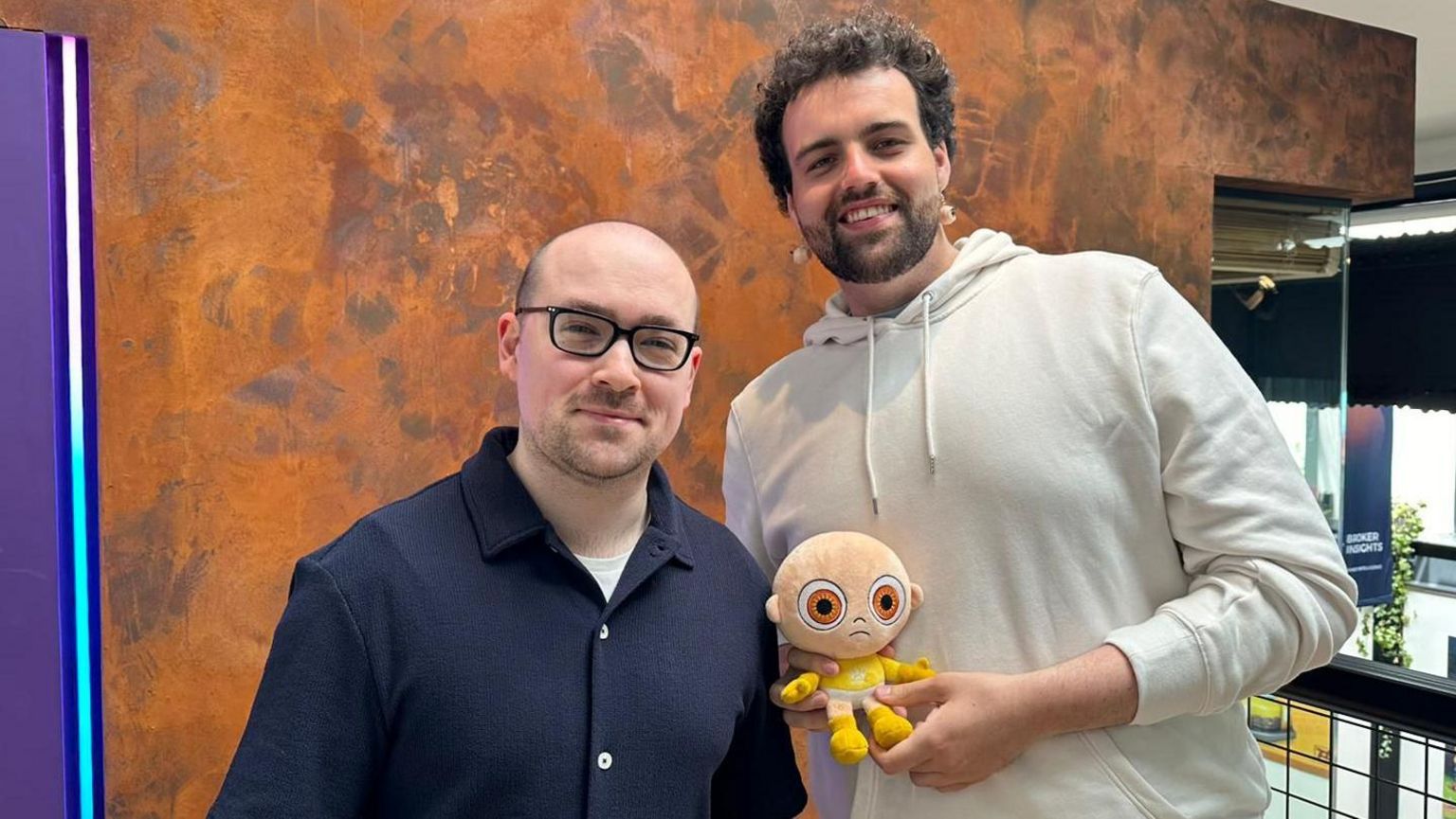 A man with glasses and a shaved head in a blue shirt stands next to a taller man with curly hair who wears a cream-coloured hoodie. Both are smiling, and the taller man is holding a baby soft toy with large yellow eyes.