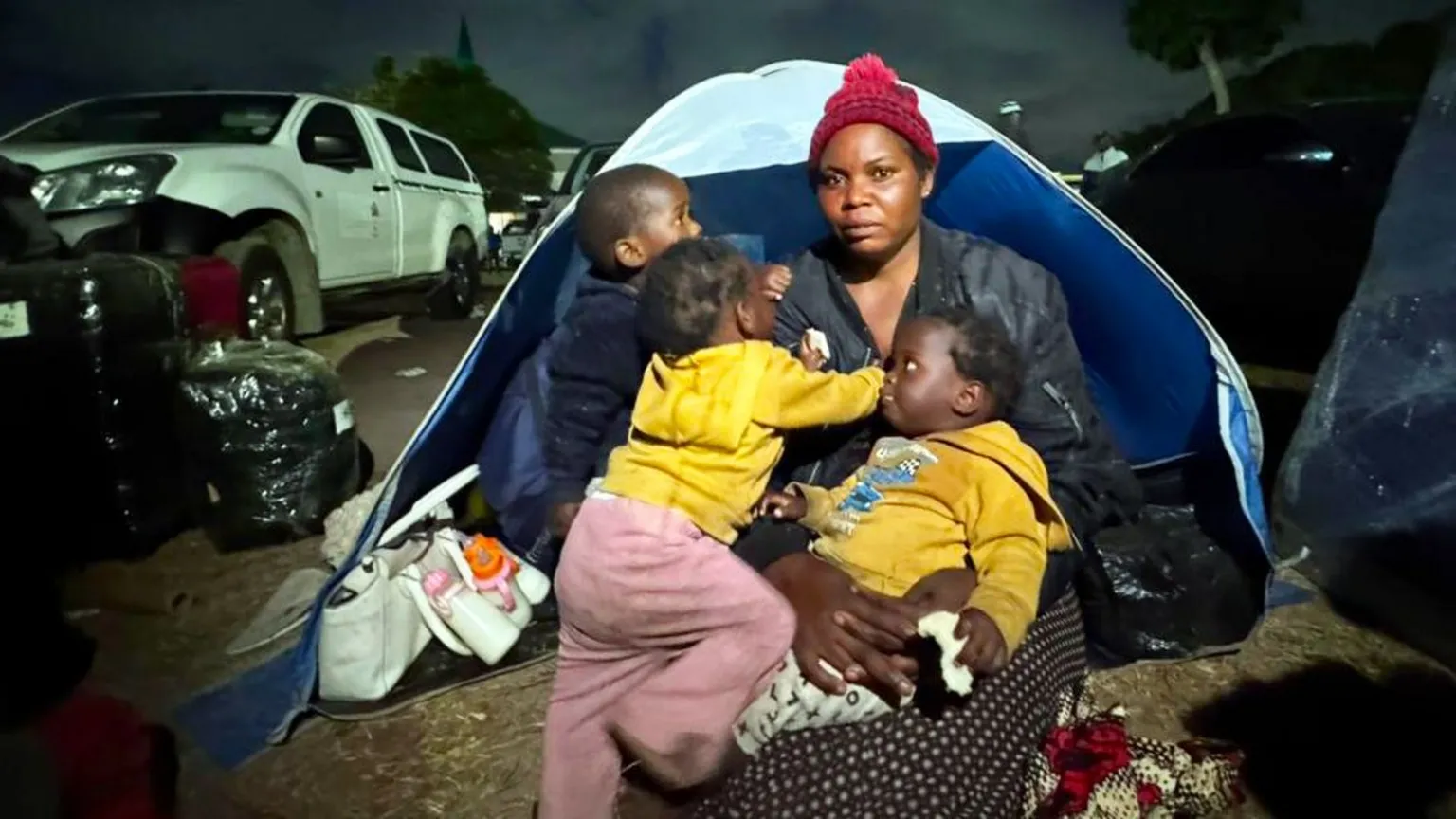 Thuthuka Zondi / BBC Esnat Joseph, wearing a maroon woollen bobble hat and black jacket, sitting in front of a tent with her baby triplets - two dressed in yellow hoodies and one black.