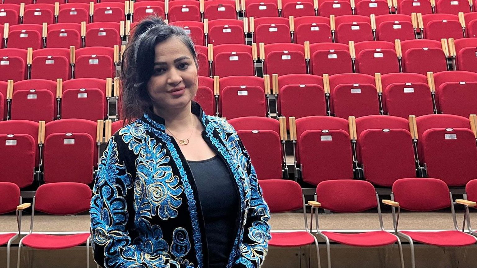 Maryam Arif wears a blue and black embroidered jacket, standing in an empty theatre