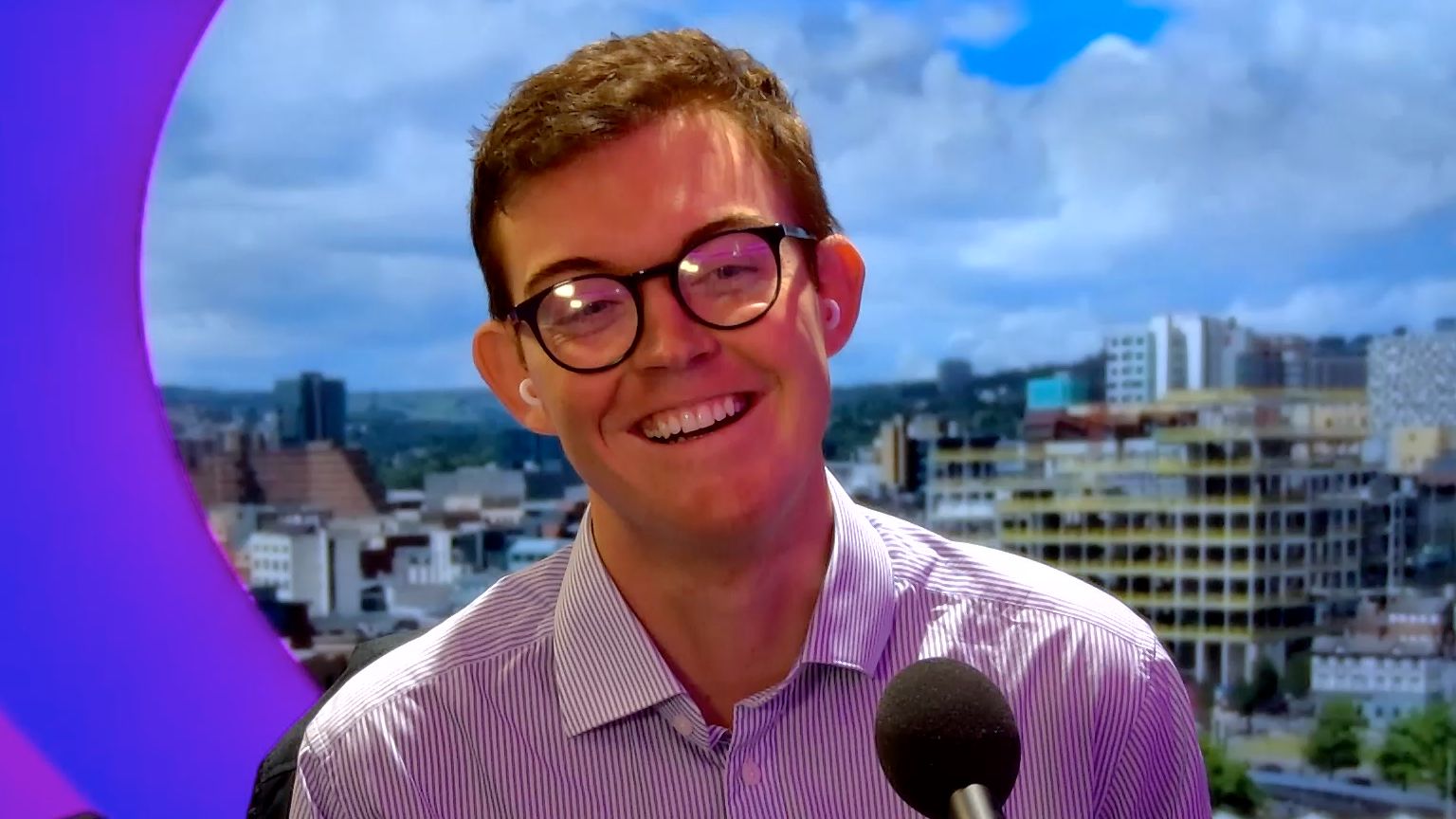 A laughing Charlie Pearson in the purple BBC Radio Sheffield studio. He sits in front of a microphone, wearing a striped shirt and round glasses. He has short brown hair.