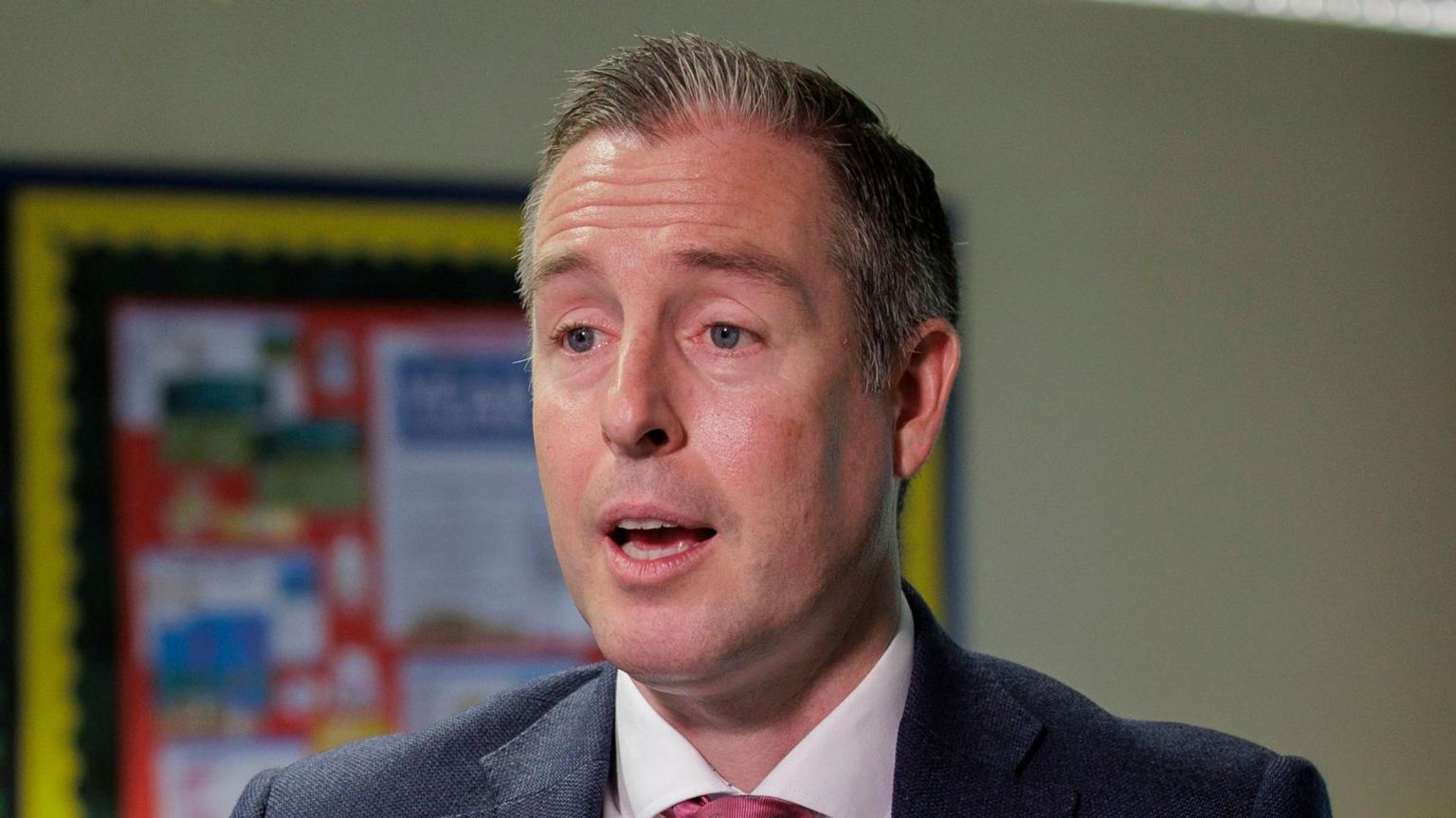 Paul Givan, wearing a dark jacket and pink tie with a white shirt