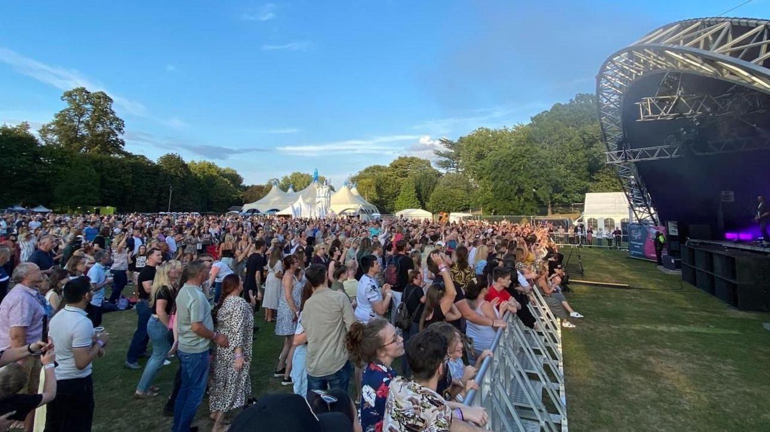 A large crowd of people standing in front of the stage in Lower Castle Park. The sky is blue and in the background are trees.