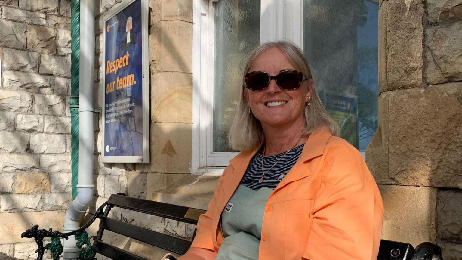 A woman with light-coloured hair and an orange jacket sitting on a bench at a railway station, smiling.