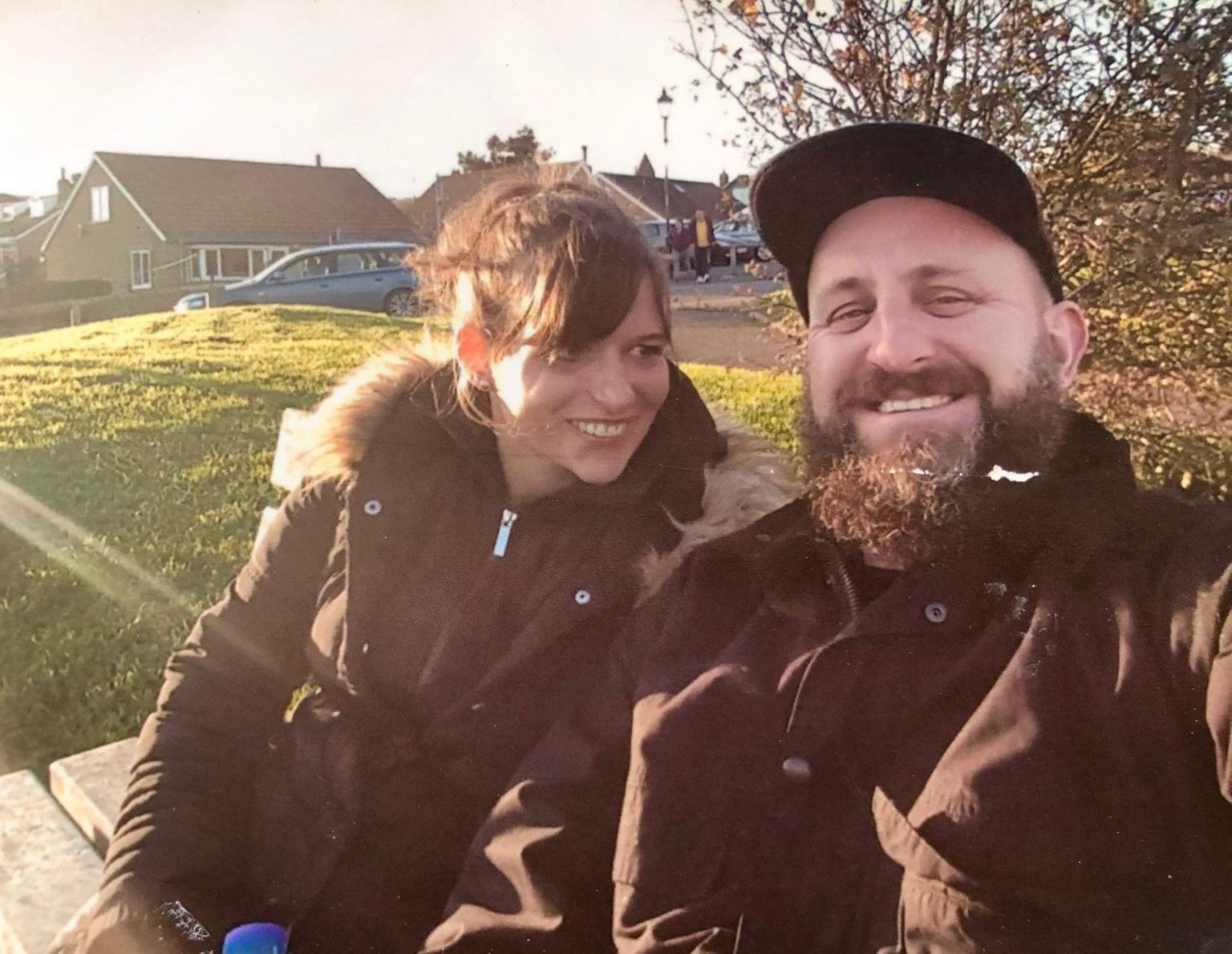 Aymie is pictured sitting on a bench outside in a winter coat looking at her partner Paul who smiles at the camera 