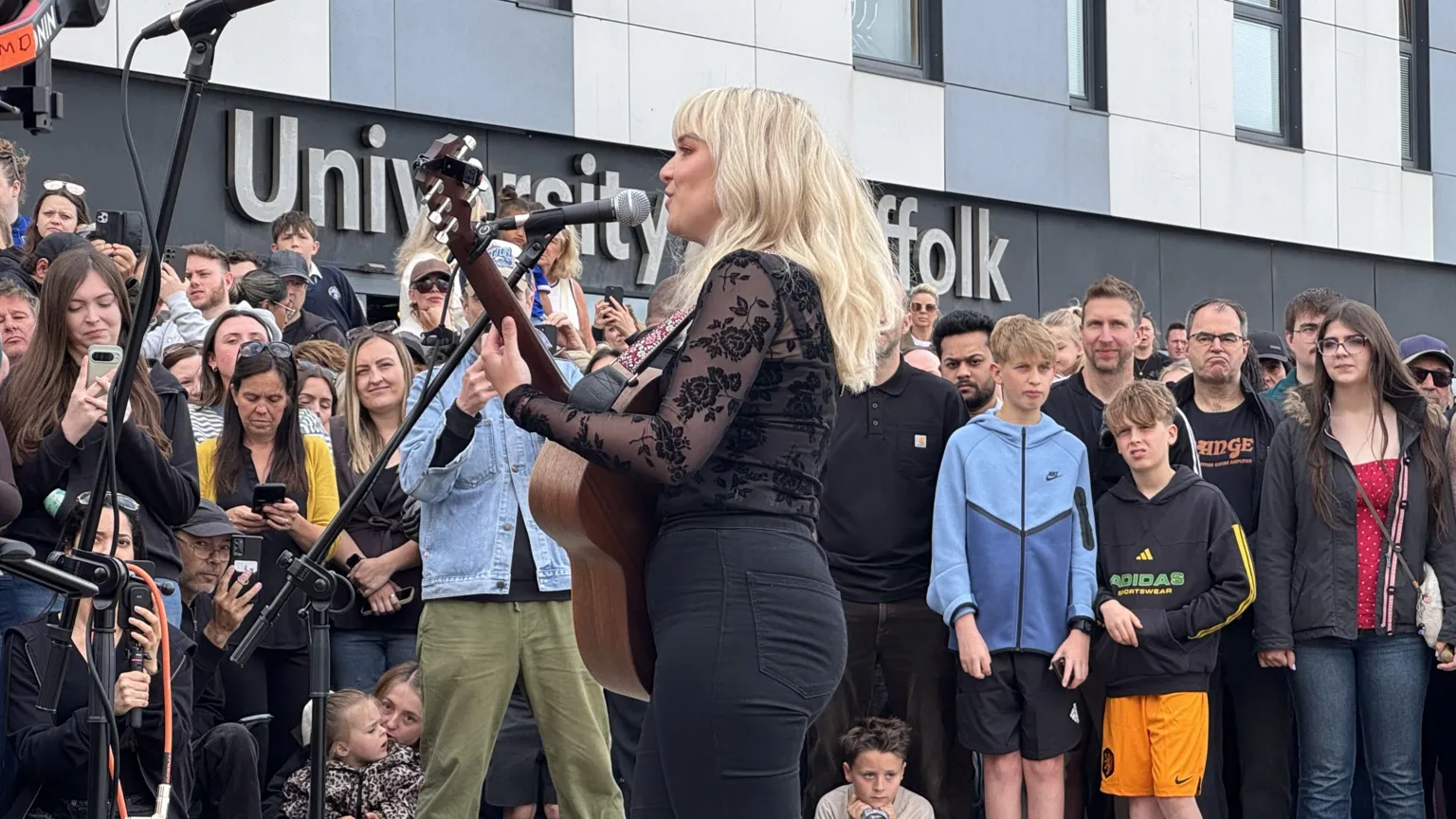 Zoie O'Brien/BBC A woman with long blonde hair wearing a black top and black jeans plays a guitar as she sings into a microphone. A crowd of people watch her, with some of them taking photos.