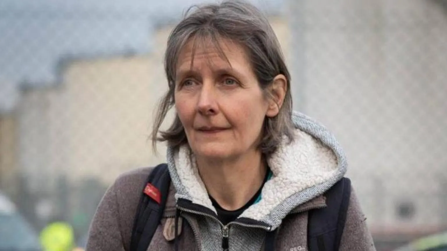 The image shows a middle-aged woman with shoulder-length brown hair wearing a gray hooded sweatshirt over a dark jacket. She appears to be outdoors with blurred buildings visible in the background. Her expression looks serious or contemplative as she faces slightly away from the camera.