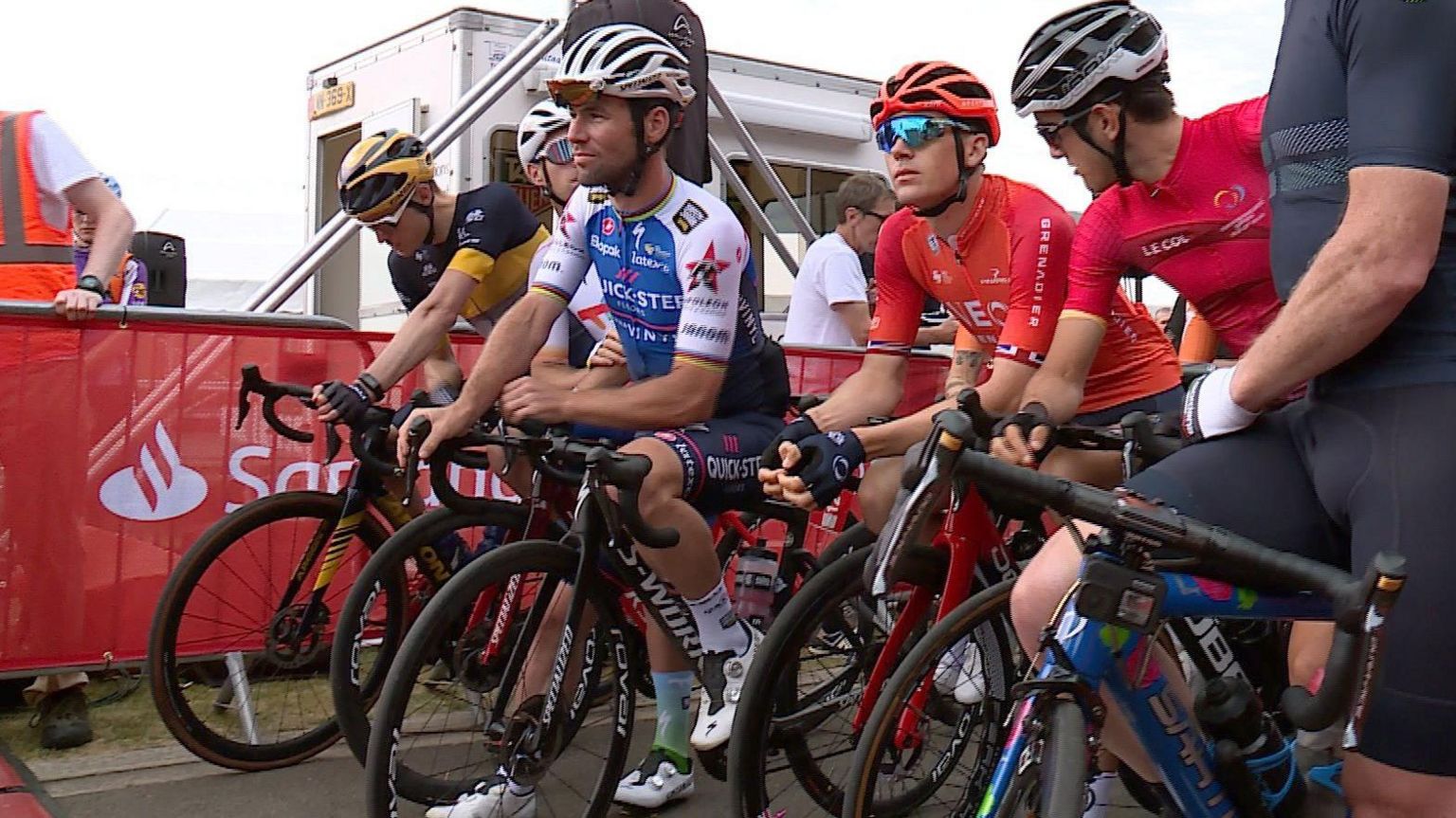 Six cyclists on bicycles at the start line of a race, wearing multicoloured lycra outfits and helmets line up at the beginning of a race. Mark Cavendish is in the middle wearing blue and white.