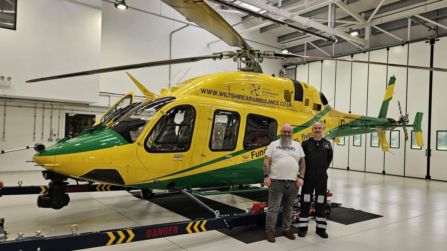 A picture shows Mr Hornsey inside the Wiltshire Air Ambulance base, next to a member of the charity's crew. They are standing next to a yellow and green helicopter with the charity branding on it, and are smiling at the camera. 