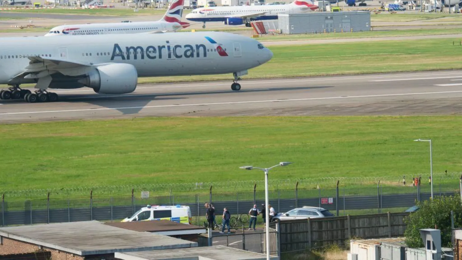 The image shows an American Airlines aircraft on a runway at what appears to be Heathrow Airport. In the foreground, several people and vehicles including a white van are gathered near airport perimeter fencing. British Airways planes are visible in the background on the tarmac.