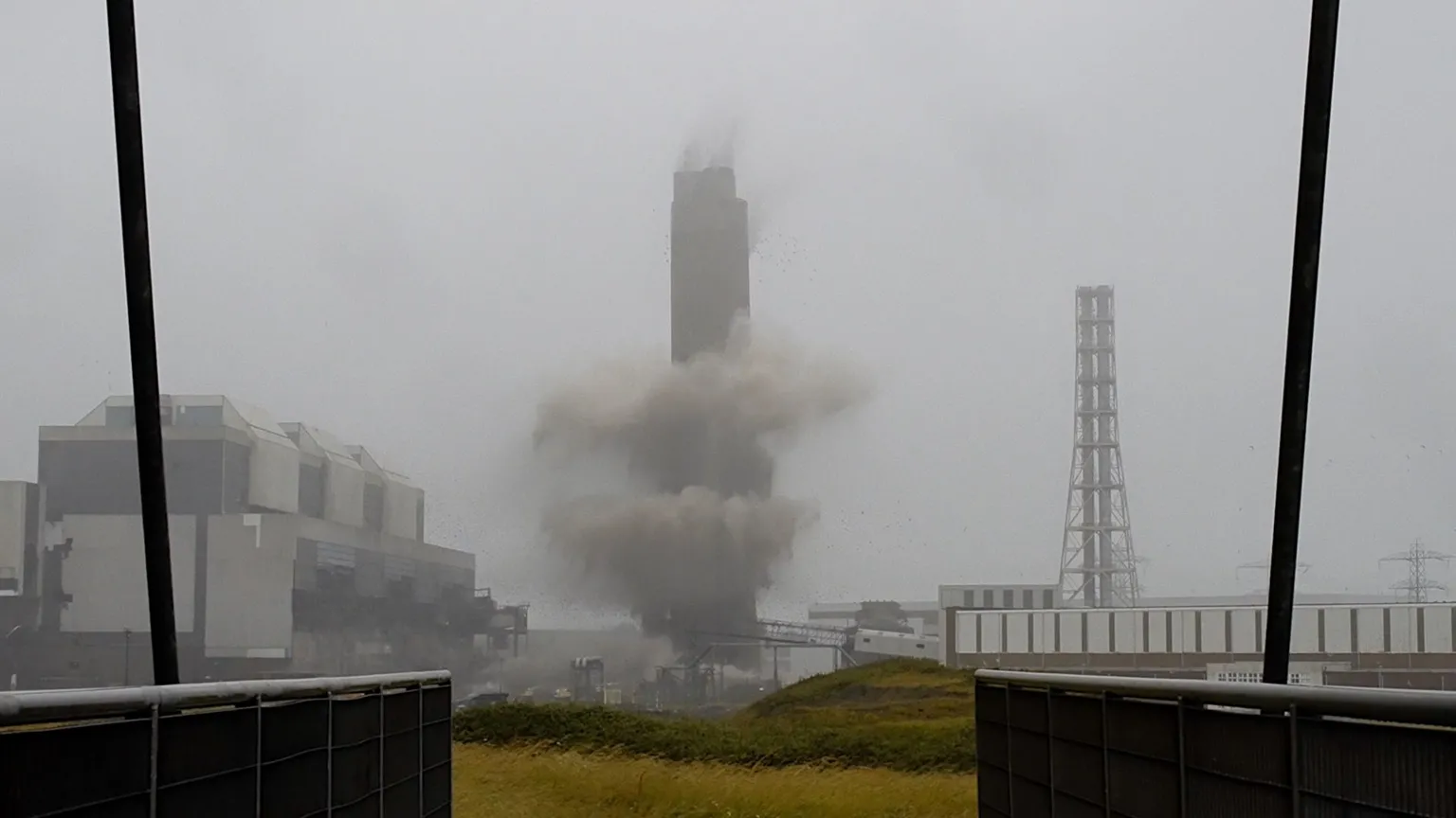 Smoke surrounds the a tall chimney as it is demolished. It is a grey, foggy day with a white sky behind the chimney. A building can be seen to its left-hand side.