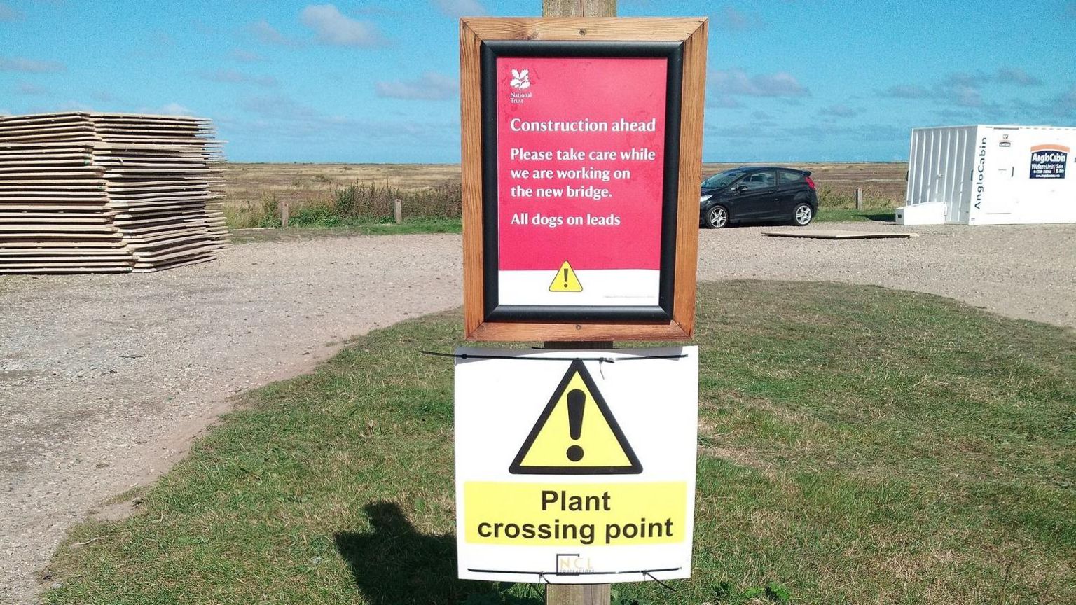 Two warning signs at the site of the new bridge. One reads: "Plant crossing point". There is a patch of grass next to a path with a car and a field in the distance.