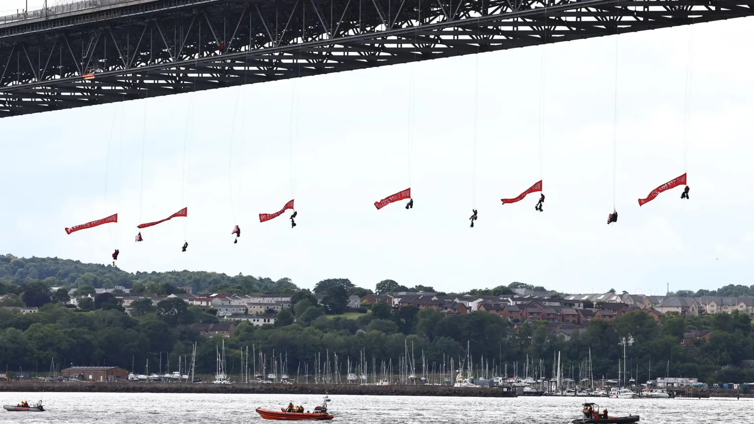The image shows several protesters suspended from red banners beneath a large steel bridge structure. Below, there's a marina with boats and residential buildings on hillsides. Small boats are visible in the water, likely monitoring the situation. The protesters appear to be abseiling from the bridge.