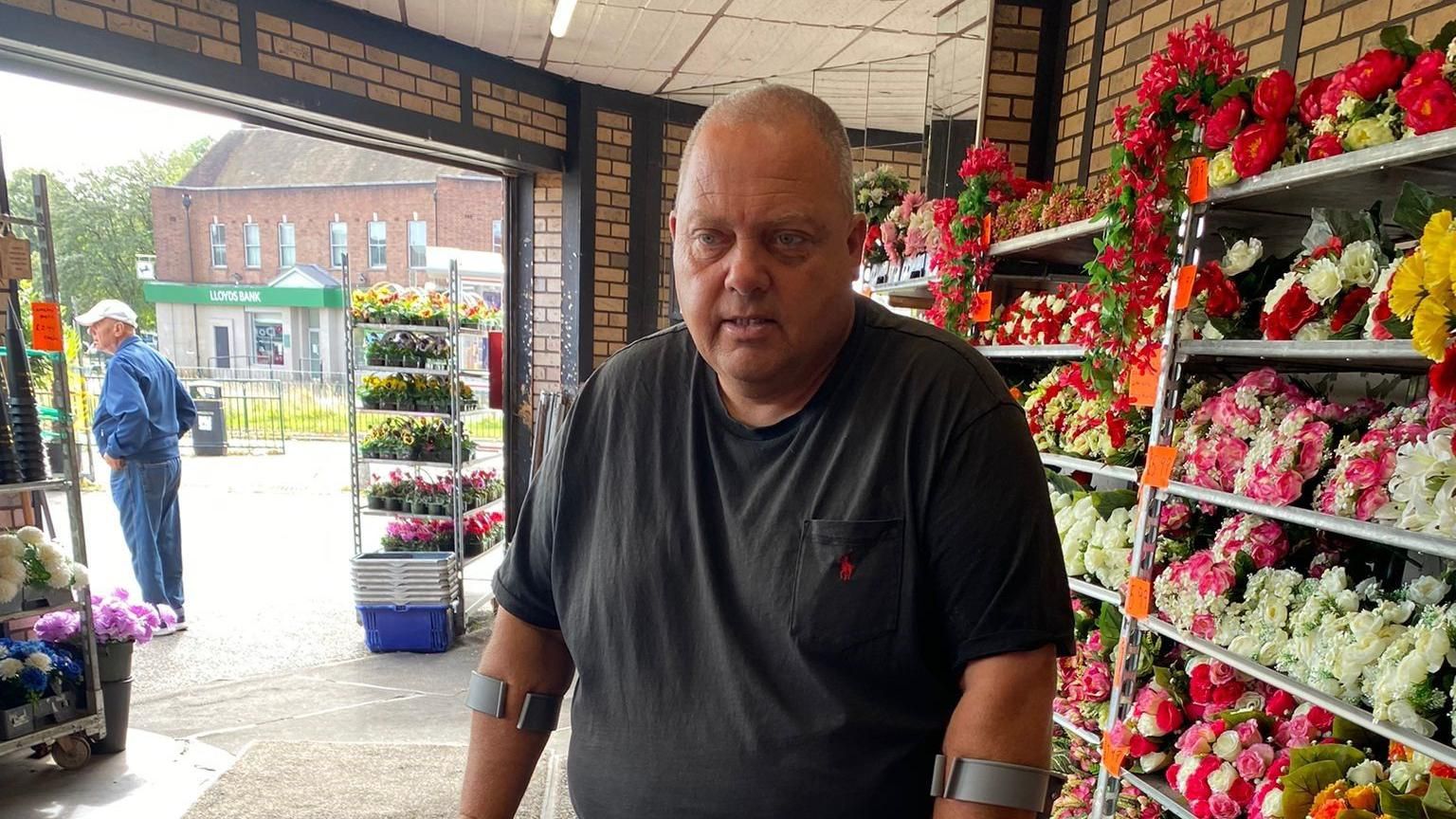 A man in a black t-shirt and using crutches stands in front of rows of colourful flowers in a shop