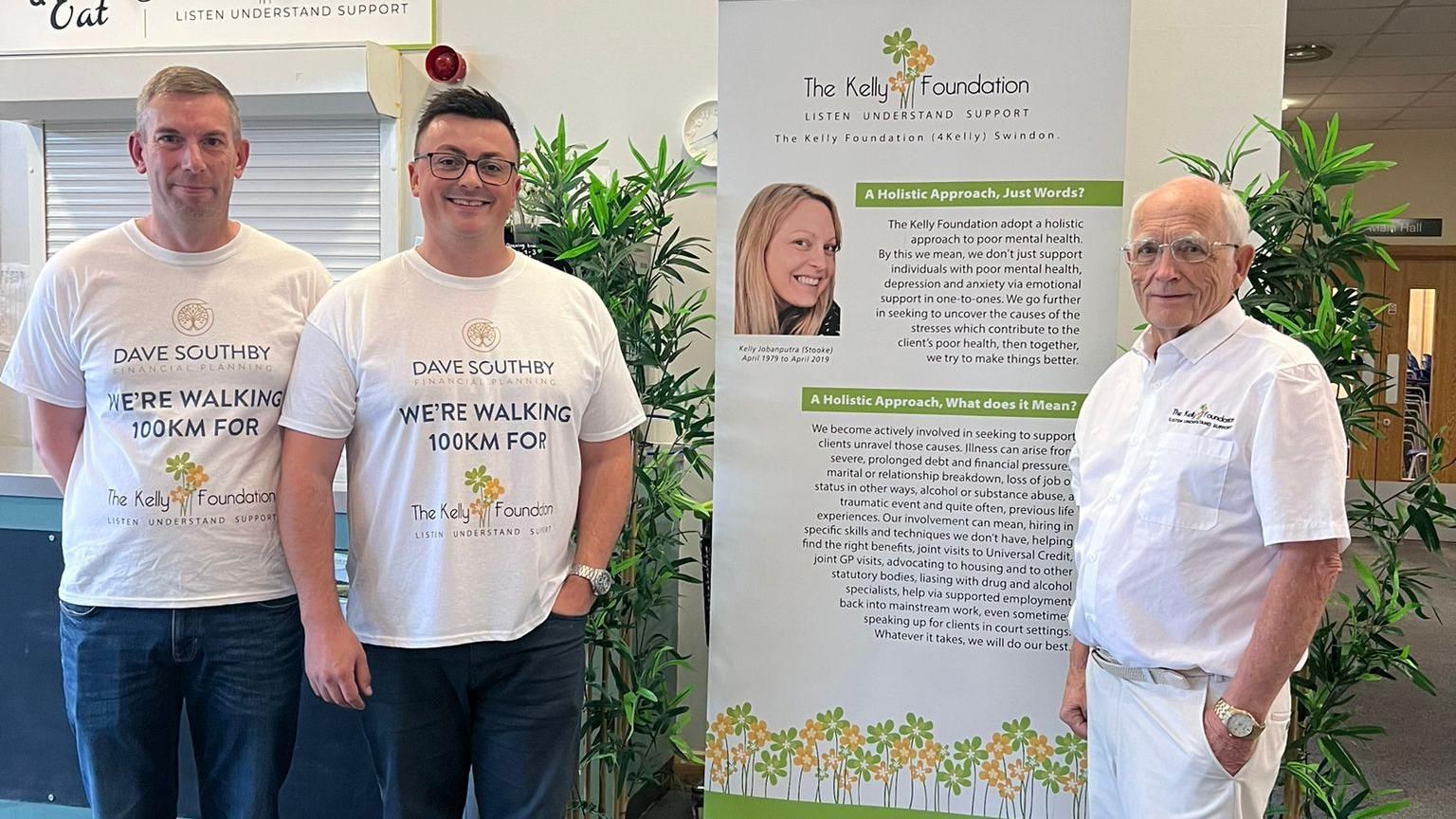 Dave Southby and his colleague Neil Sawyer posing beside a Kelly Foundation banner. They are both wearing white t-shirts which say 'Dave Southby Financial Planning. We're walking 100km for the Kelly Foundation.'
On the other side of the banner, charity chairman John Stooke is also looking at the camera wearing all white.
