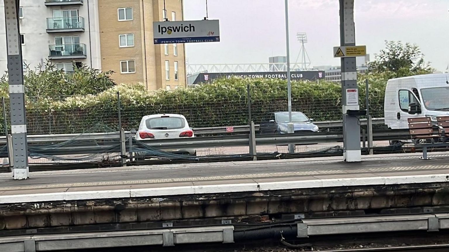 An empty platform at Ipswich railway station with an Ipswich sign above the platform, which backs onto a car park with two cars and a van in and Ipswich Town FC ground in the background