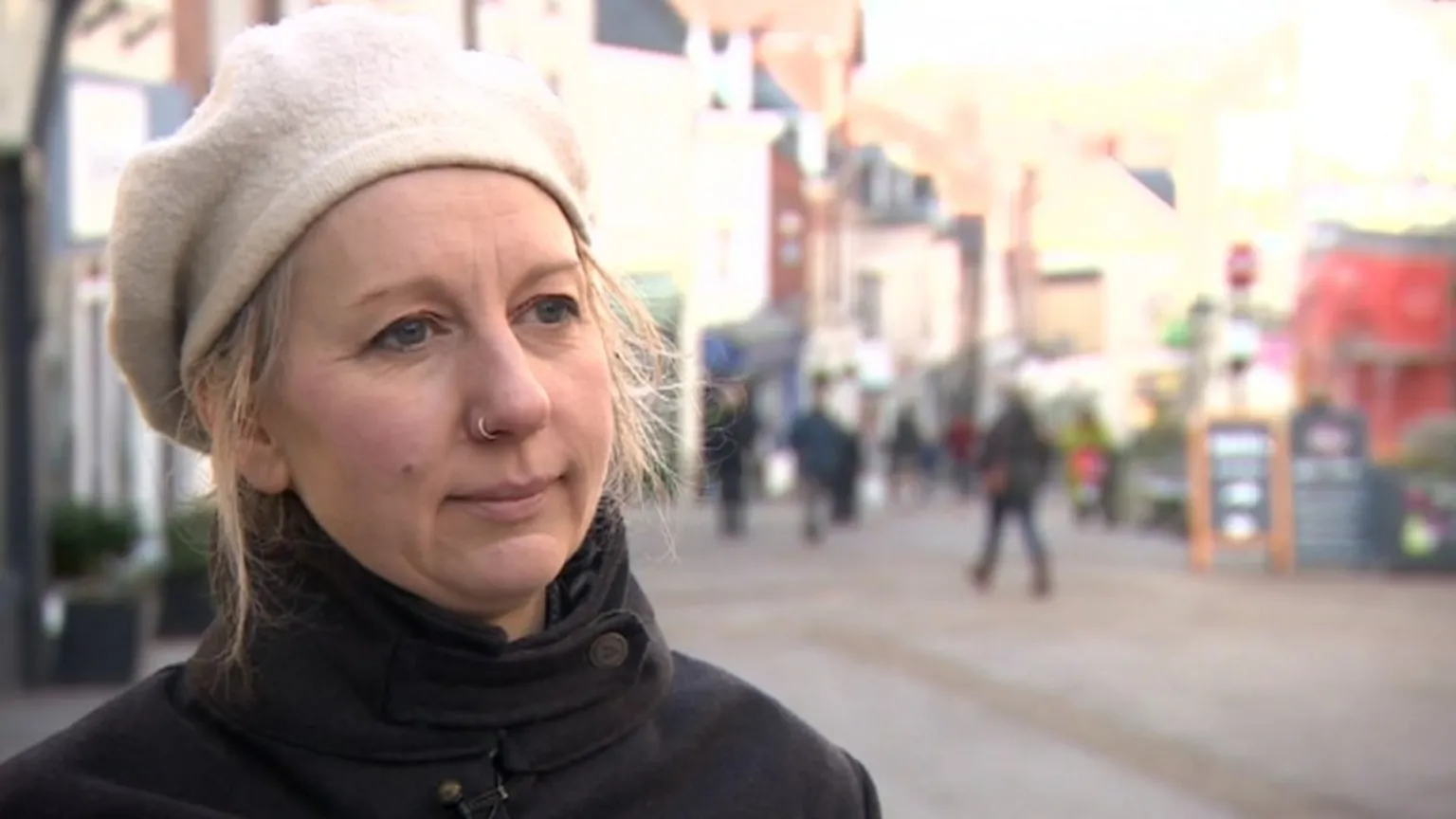The image shows a woman with blonde hair wearing a light-colored knit beanie and dark jacket, standing on what appears to be a pedestrian street or plaza. She has a nose piercing and is looking directly at the camera with a slight smile. The background shows a blurred urban setting with buildings and people walking.