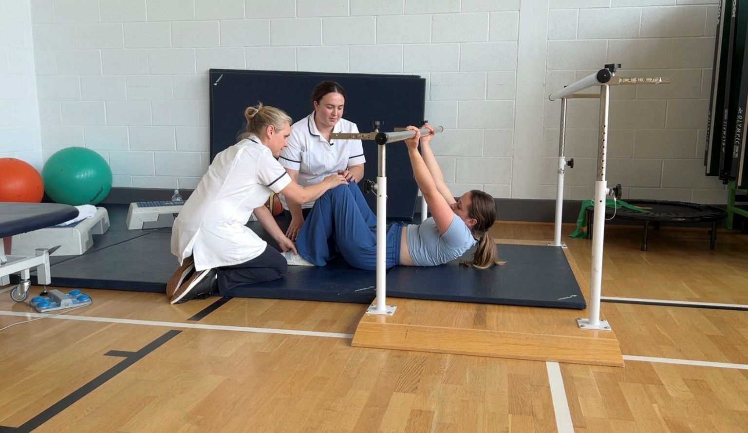 Saffron Cresswell is lying on an exercise mat during a rehabilitation session supported by two women both wearing white nursing uniforms