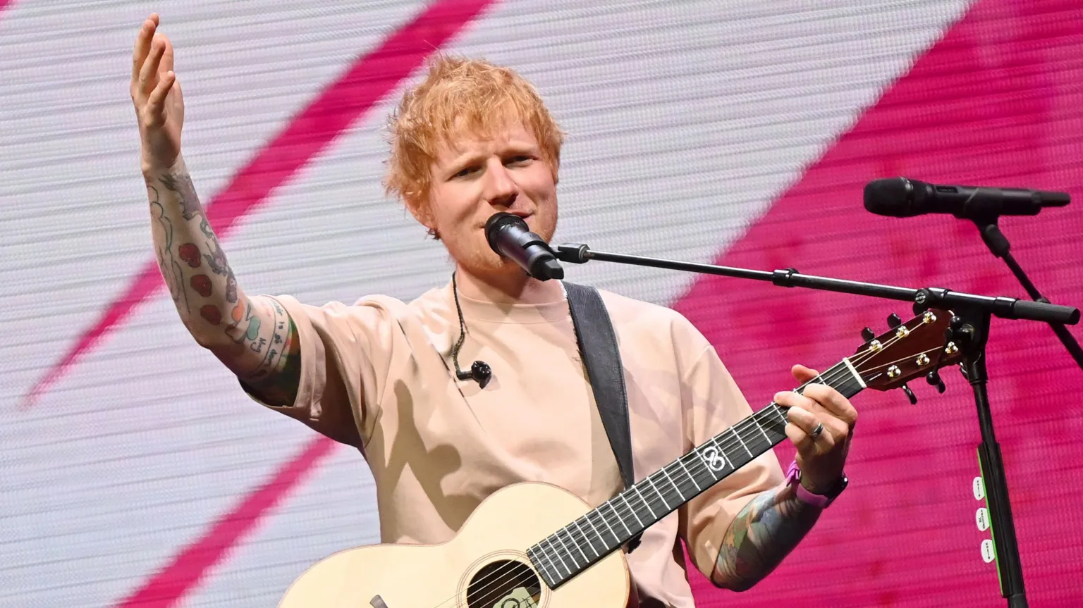 Getty Images Ed Sheeran singing into a microphone on stage with one hand on the neck of his guitar and the other raised.