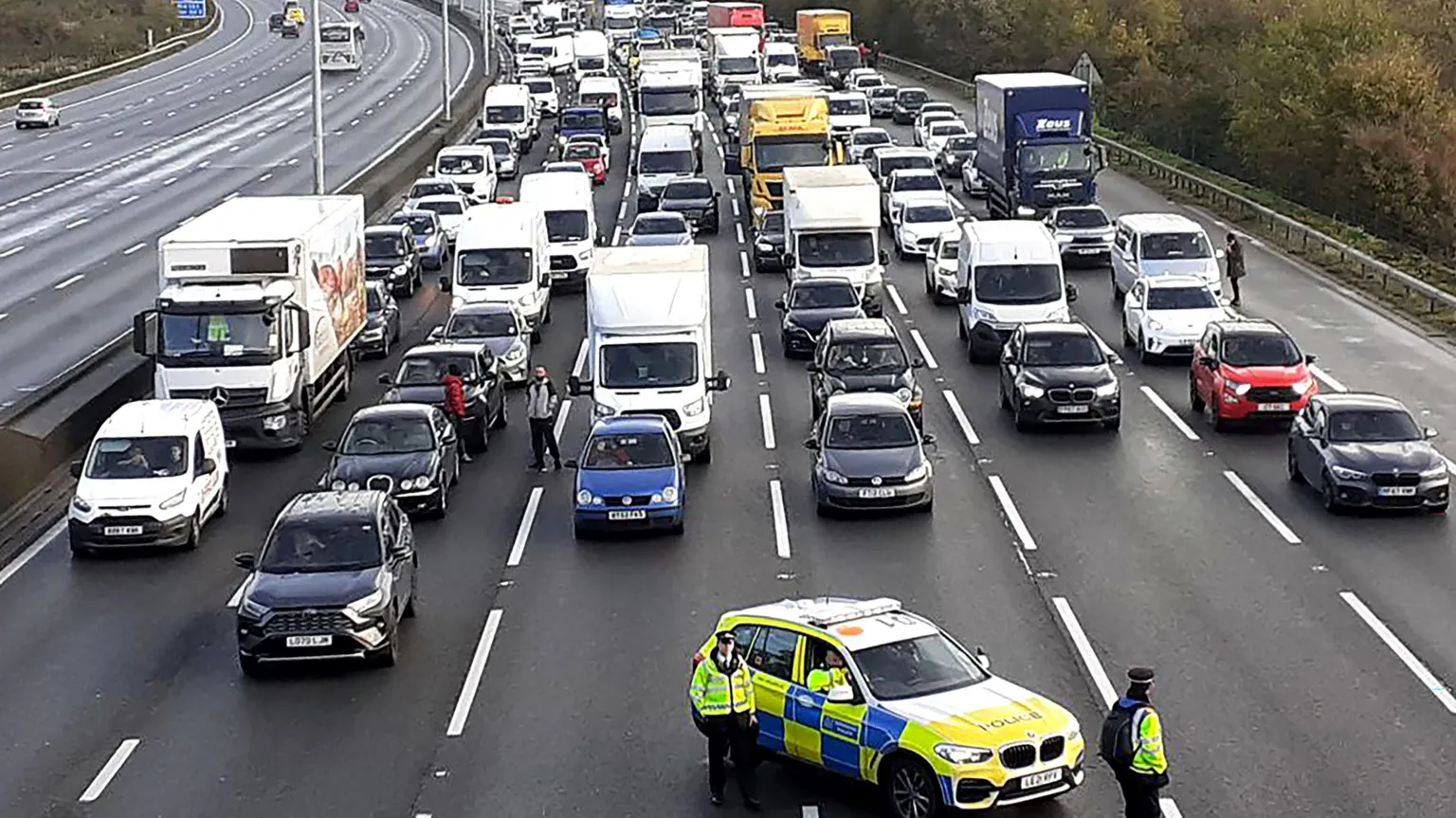 The image shows heavy traffic congestion on the M25 motorway with vehicles at a complete standstill across multiple lanes. A police car with high-visibility markings is positioned on the road with an officer standing nearby. The scene depicts the traffic disruption from environmental protests in November 2022.