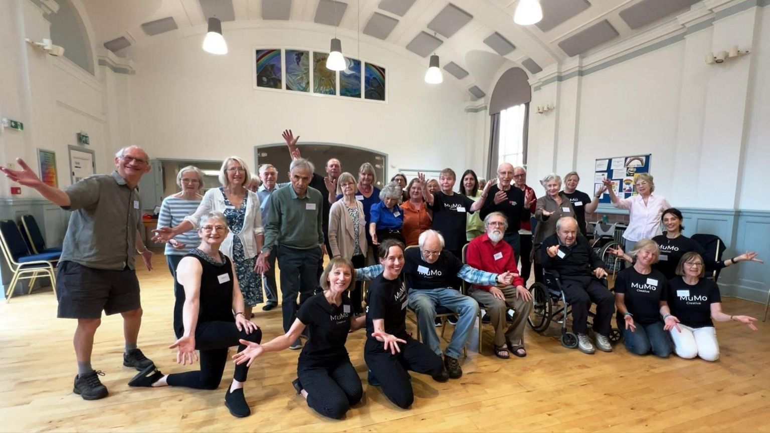 MuMo Creative with a crowd of elderly people in a hall, all of the group are posing for the camera with their arms outstretched in a dance pose.