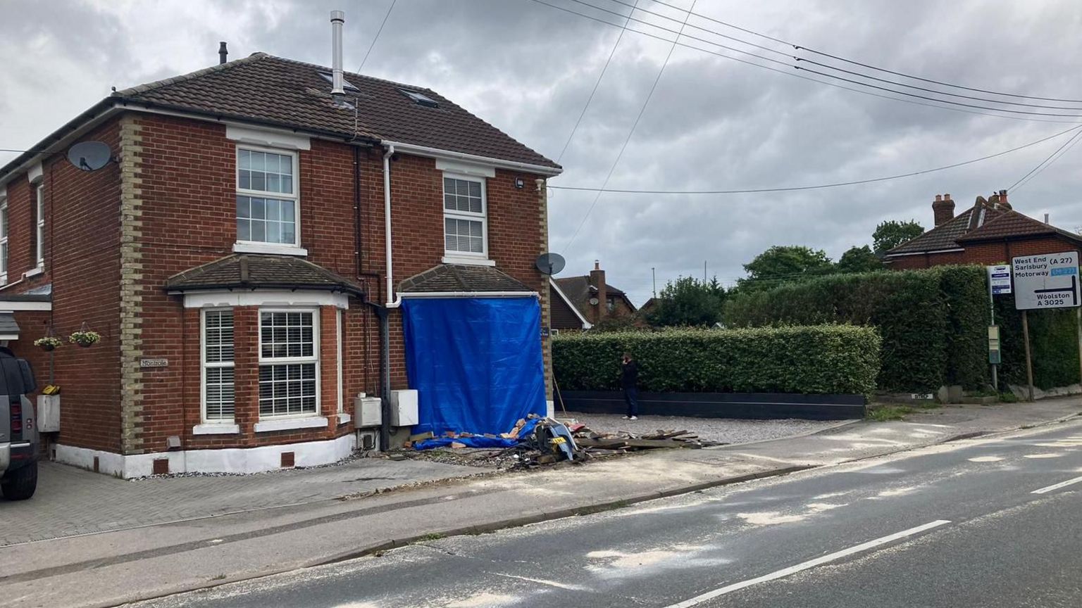 A red brick brick semi-detached house with a blue plastic covering over the bottom right bay window with wreckage scattered outside