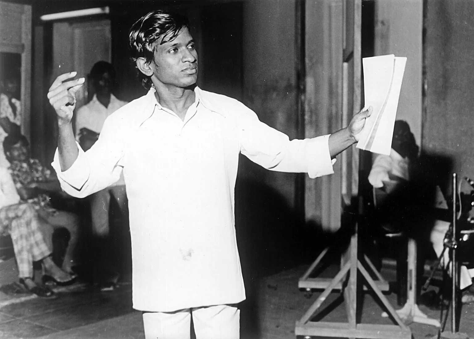 Ananda Vikatan Ilaiyaraaja stands holding a white sheet of paper as people sit in the background and watch him