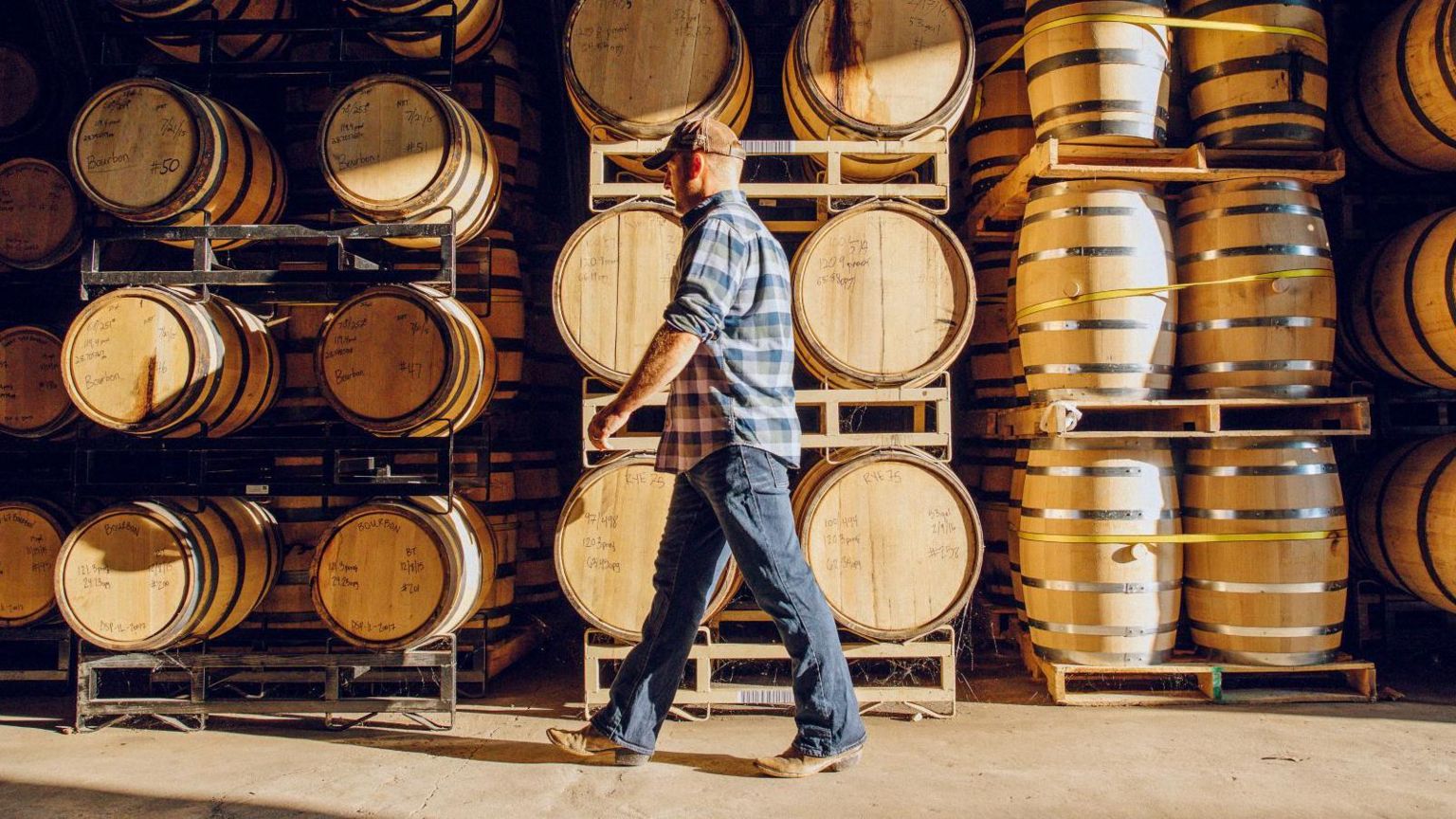Man in blue checkered shirt, jeans and baseball cap walks by a wall of stacked whisky barrels in a distillery 