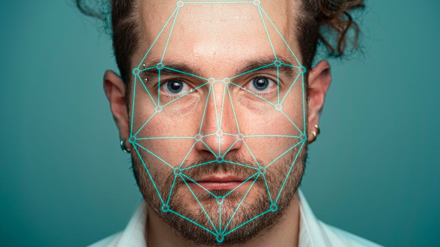 Close up of a man with blue eyes, a dark, groomed bead and his hair is back off his face. He has an eyebrow piercing and is wearing hooped earrings. The background is teal. There are a series of green lines running across his face.