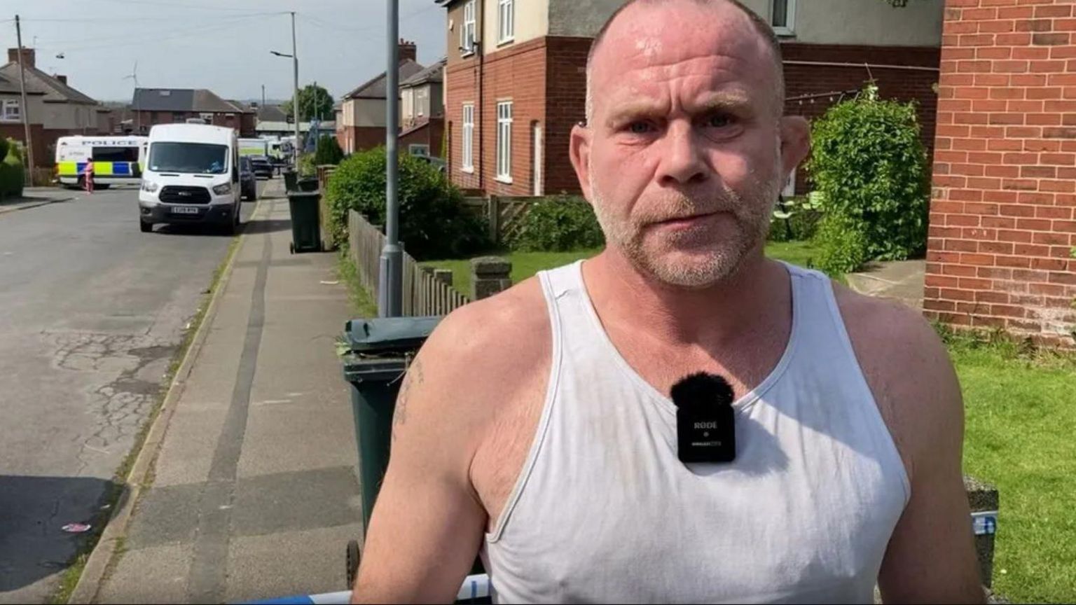 A picture from a previous interview with Lee Crisp that shows him in a tank top in front of a police cordon. He is nearly bald and has grey stubble. There appears to be a tattoo on his right upper arm.