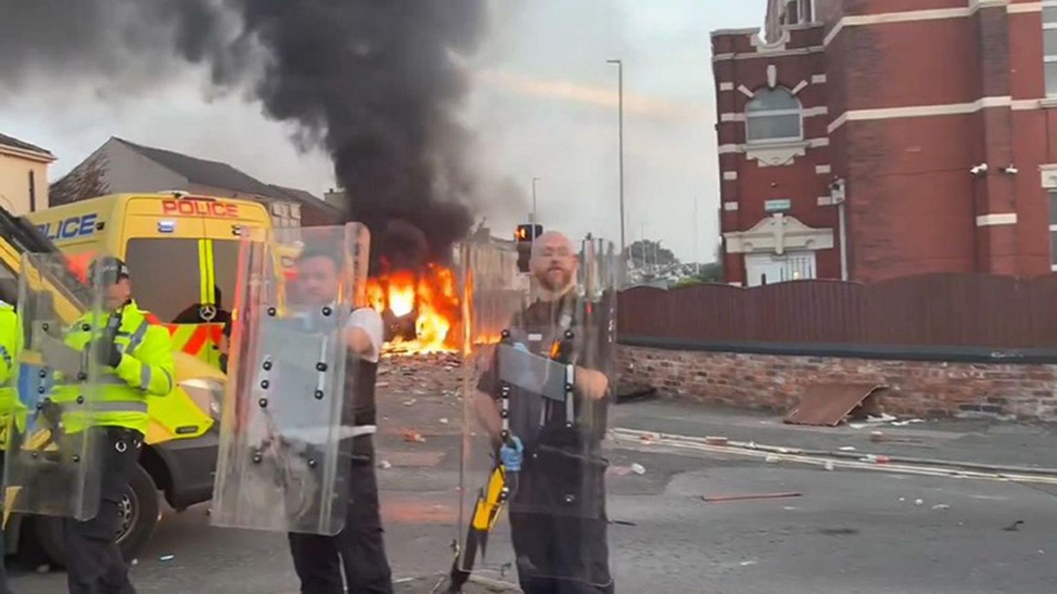 Police hold in riot shields stand in front of a red brick building. In the background a police van is on fire.