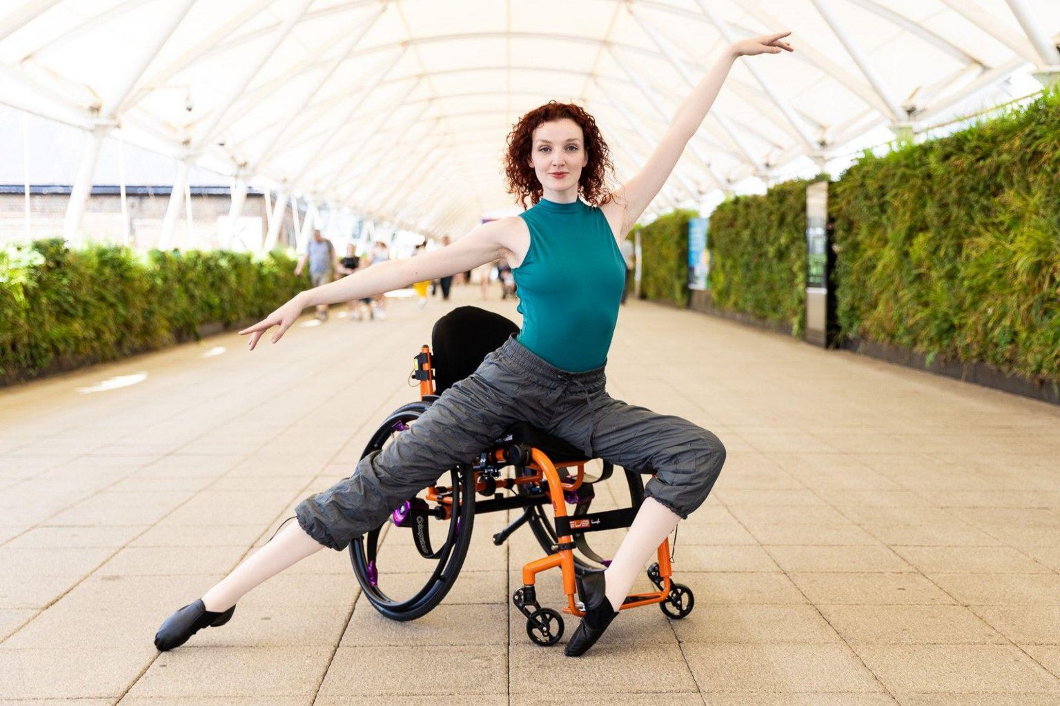 Kate Stanforth in a ballet pose perched on an orange wheelchair, wearing a green leotard and grey trousers. 