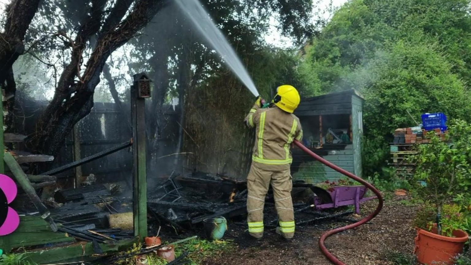 A firefighter with a hosepipe shooting water up at a tree which appears to be black from burning and the floor beneath it is also black with damaged garden items in a pile