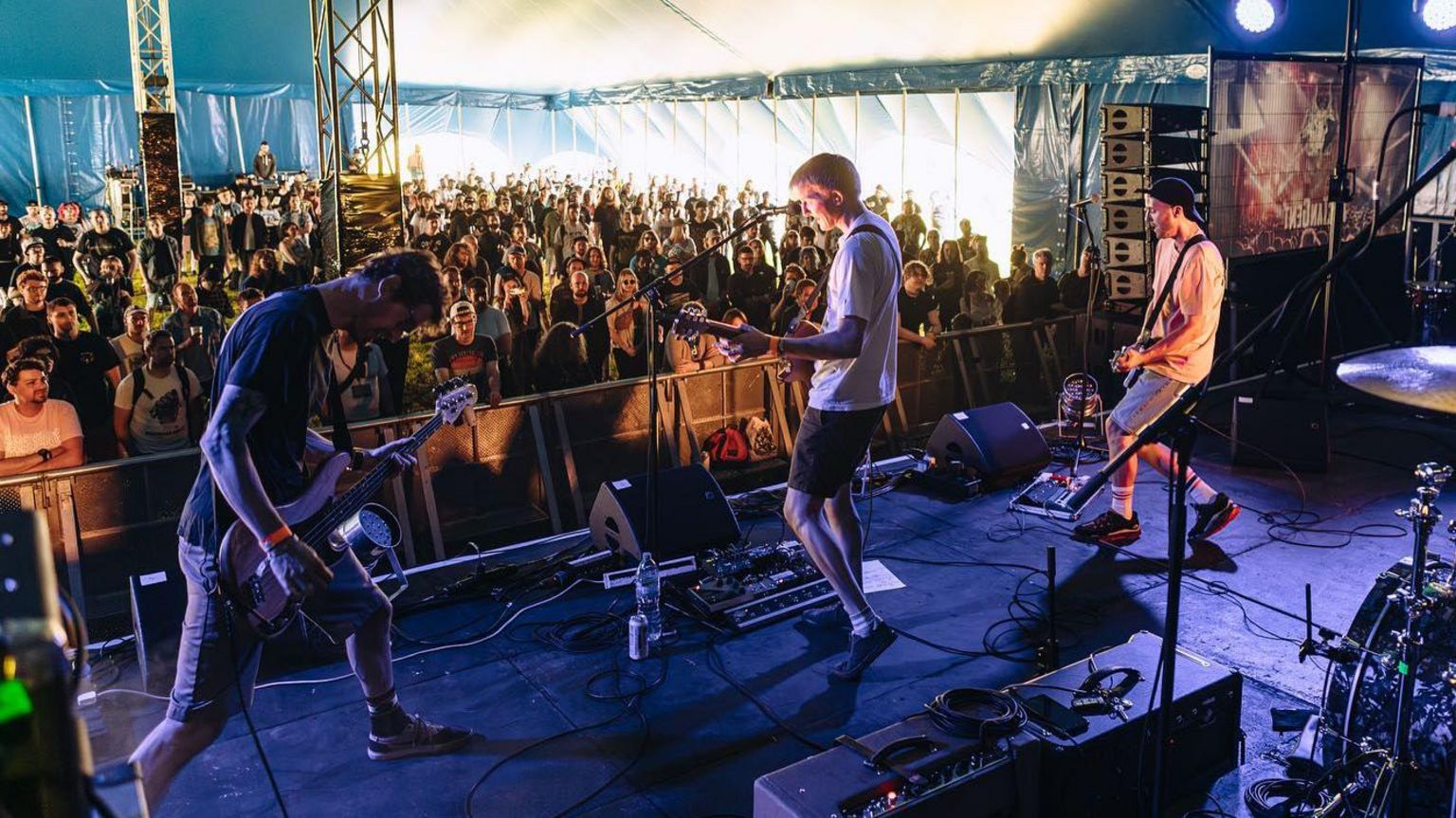 Three men on stage playing guitars and singing with a crowd of people watching