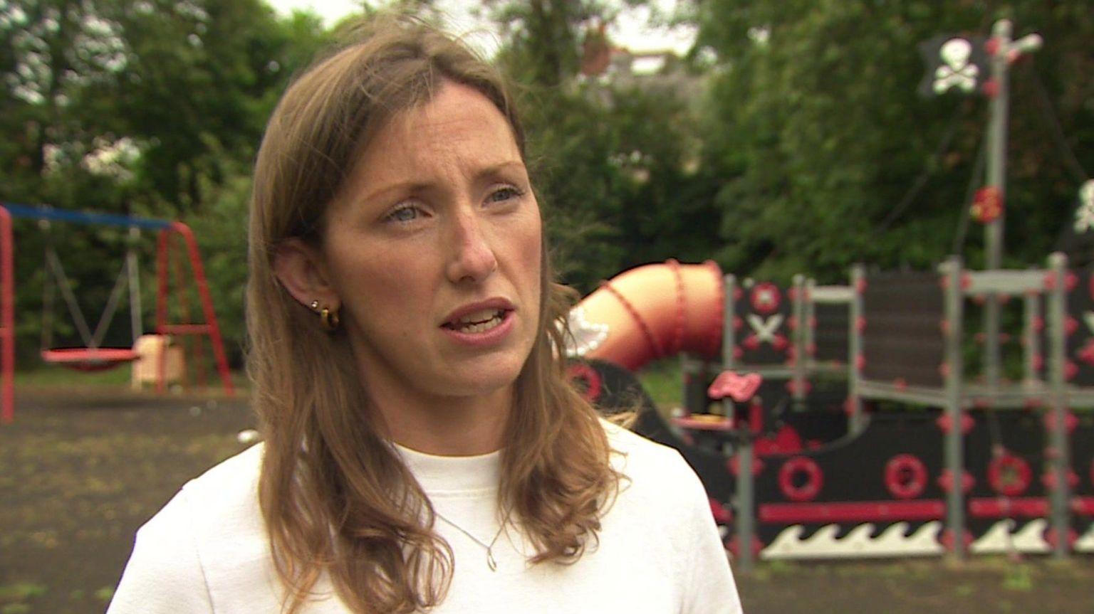 A head-and-shoulders shot of a woman with mid-length brown hair wearing a white top. She is standing in a playground and there are climbing frames behind her.