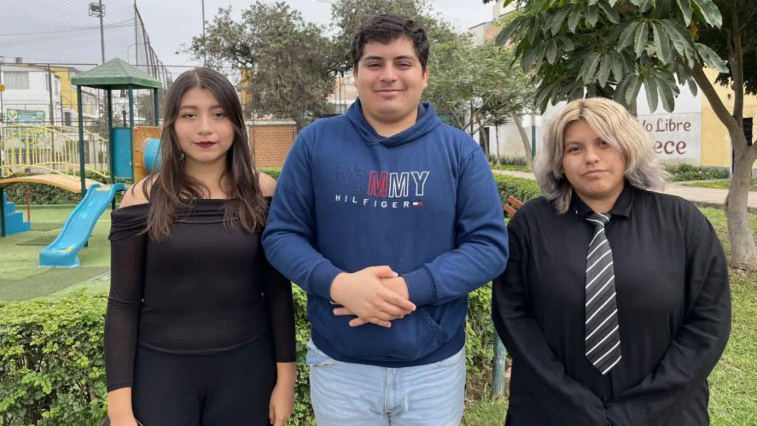 Consuelo dressed in black, next to Alvaro in a blue top and jeans and Cielo in a black top and tie. They are standing in front of an urban backdrop.
