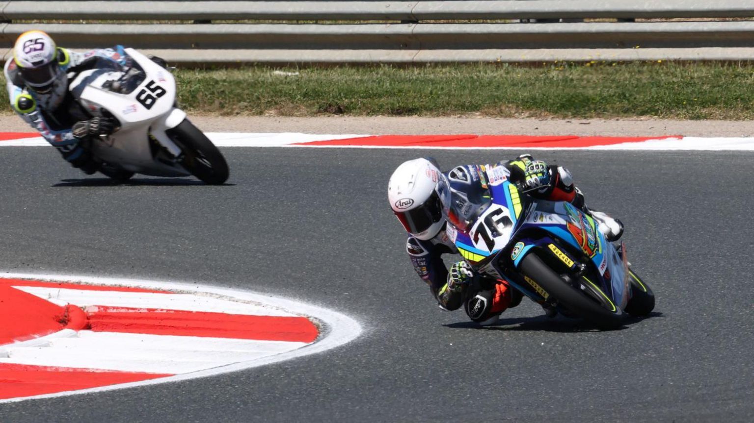 Charlie Huntingford racing his bike, which has the number 76 on it, on a track with another bike close behind him. He is wearing a race suit and white helmet. 