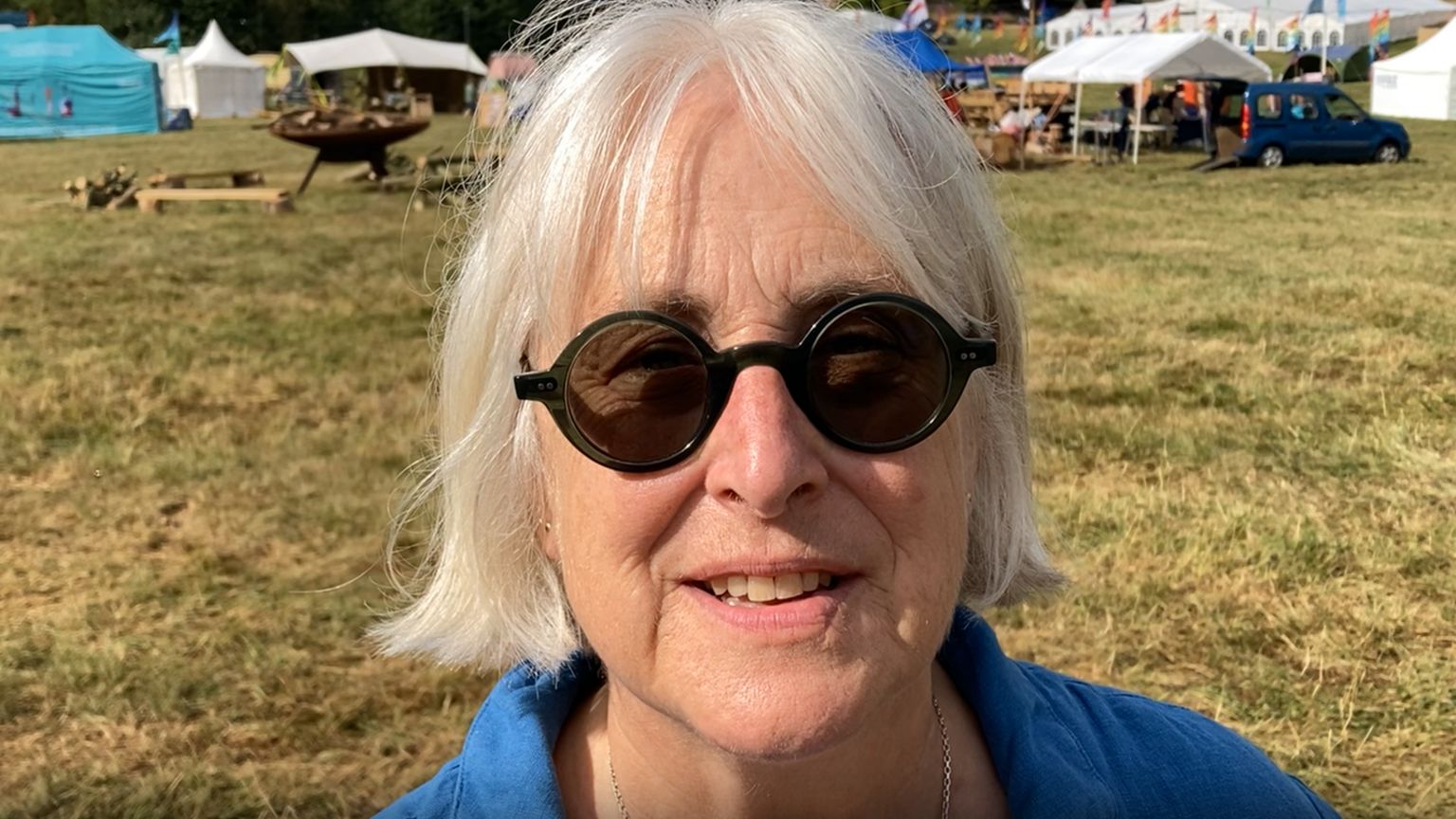 Becky Marshall-Potter photographed outside, she is wearing a blue jacket and dark sunglasses. Marquees, tents and cars are behind her. 