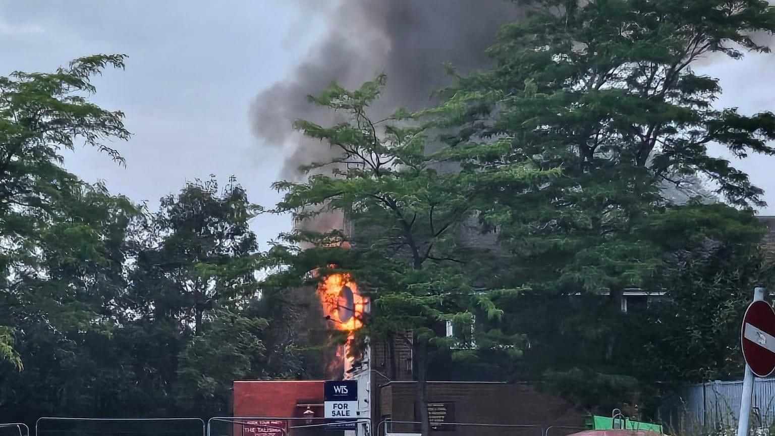 Flames flickering out from a building behind some trees. At the front of the building is a "For Sale" sign. 