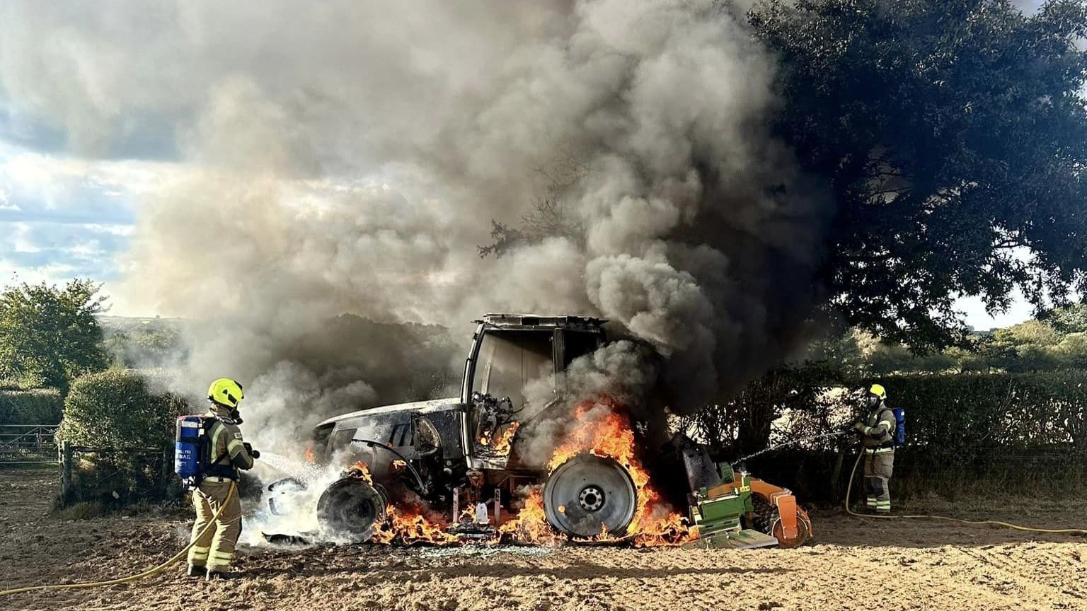 Two firefighters spray liquid onto a blazing tractor in a field. The smoke billows and flames emanate from the lower side of the tractor.