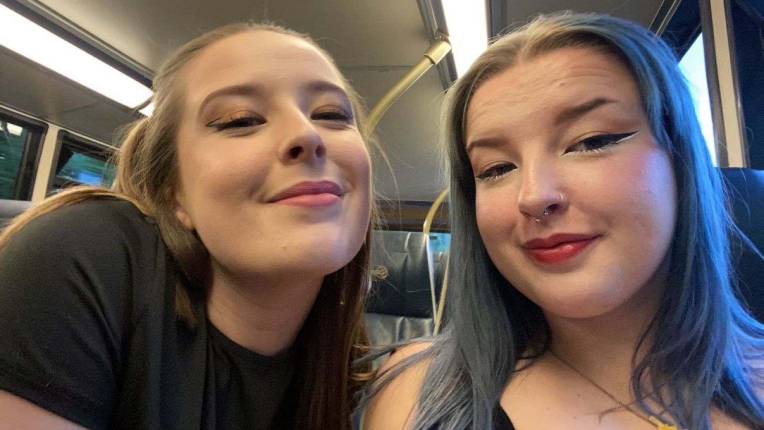 Anna and Emily sitting next to each other on a bus on their way out for the night. Anna has her curly brown hair half-up, half-down, and Emily's hair is dyed blue and worn down. Both are smiling