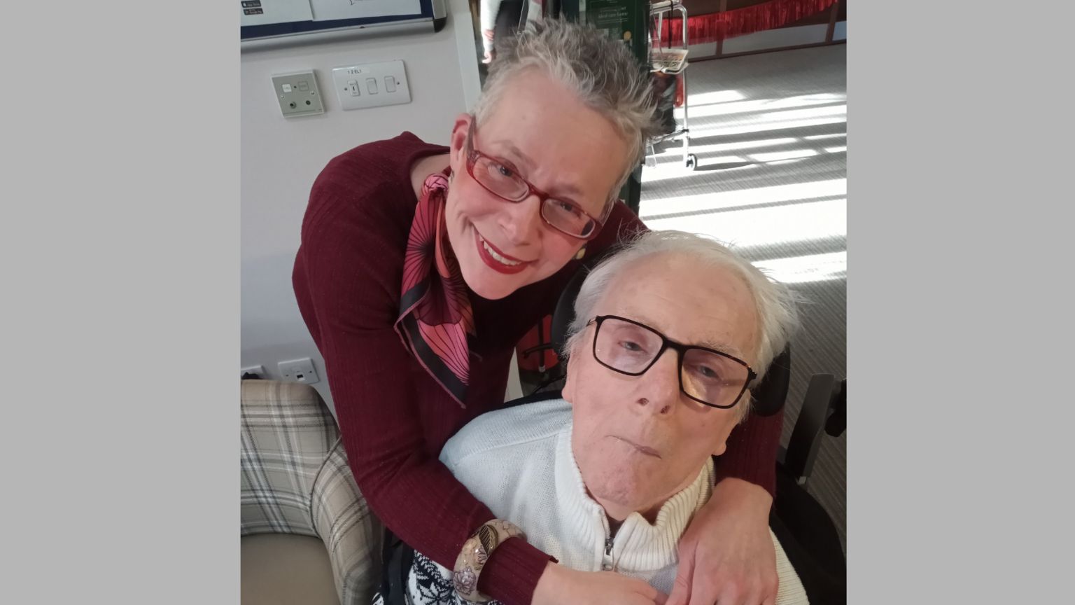 Claire Walker is pictured smiling at the camera while she hugs her father Maurice Page from behind. She is wearing a dark red jumper with a red and pink scarf and red glasses. Mr Page is pictured wearing black glasses with a white jumper.