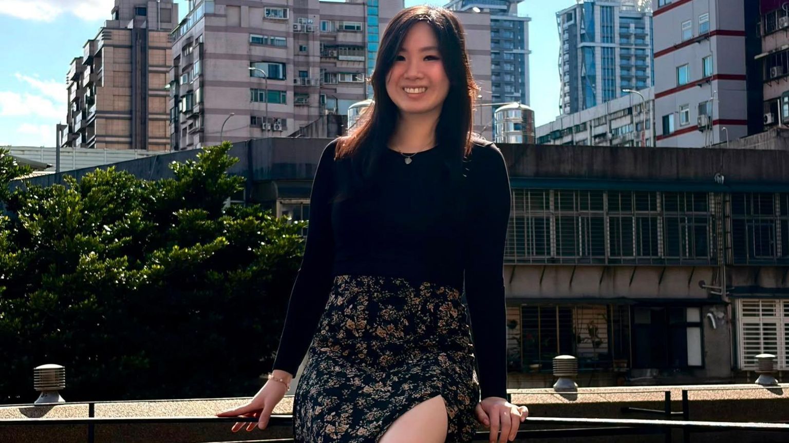 年轻投资者米歇尔·黄坐在栏杆上，在蓝天和住宅区前摆姿势拍照。